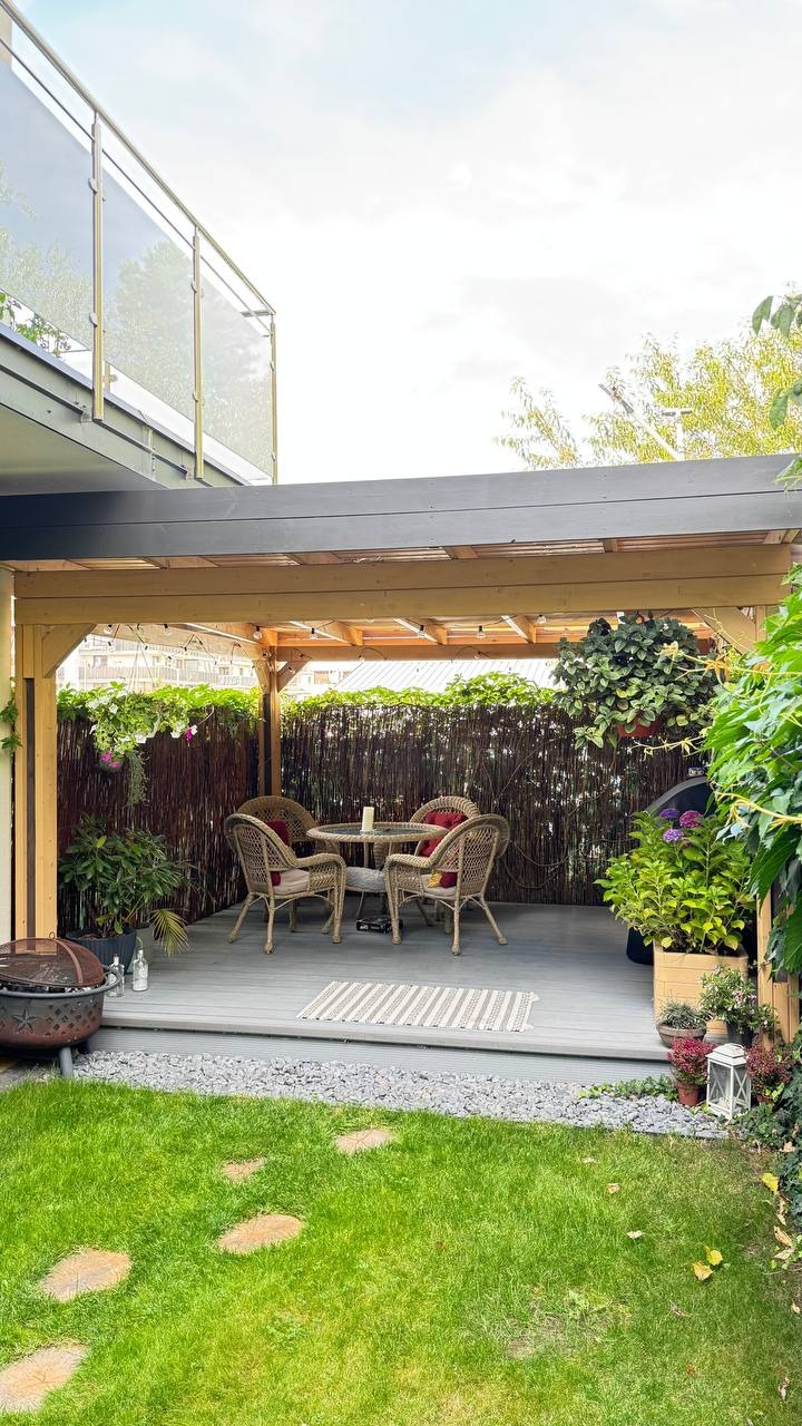 Zadaszony taras z drewnianą konstrukcją i rattanowymi meblami ogrodowymi, otoczony zielenią. Dekoracje z kwiatów w donicach i lampionów.