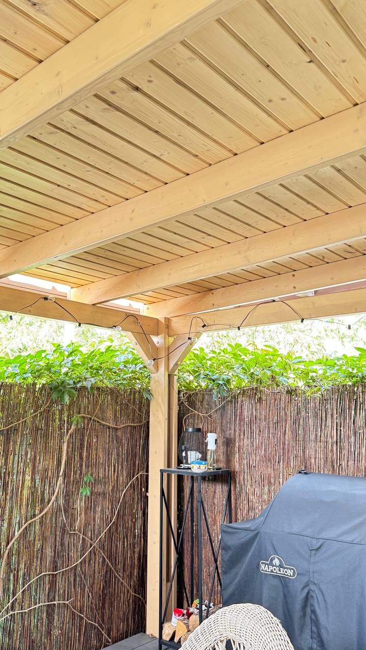 Jasna, drewniana pergola z oświetleniem girlandą, widok z dołu. W tle zielona roślinność i osłona z wikliny, obok grill Napoleon.