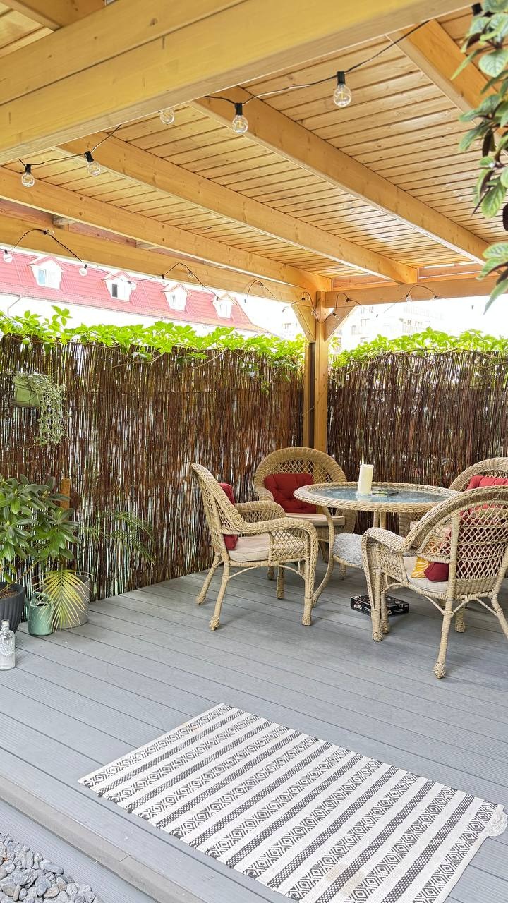 Zadaszony taras z drewnianą pergolą, rattanowymi meblami i bambusową osłoną. Dekoracyjne oświetlenie żarówkowe. Szary taras z dywanem.