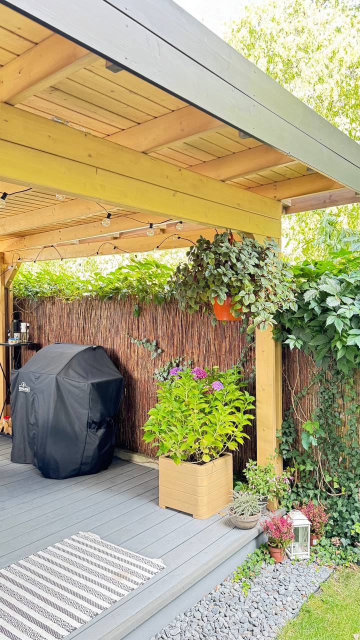 Zadaszony taras z drewnianą pergolą, oświetleniem i roślinami w donicach. Grill pod pokrowcem, mata na podłodze, kamyki dekoracyjne. Urokliwe miejsce do relaksu.