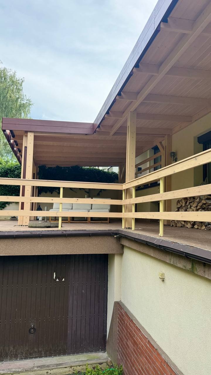 Drewniana konstrukcja tarasowa z jasnymi balustradami, częściowo zadaszona, z widocznym miejscem do siedzenia i stosem drewna w tle. Całość w naturalnej kolorystyce.