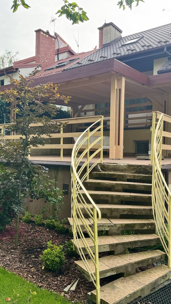 Nowoczesny taras z drewnianą konstrukcją i spiralnymi schodami w kolorze żółtym, prowadzącymi z ogrodu. Tło stanowi dom z ceglanym kominem.