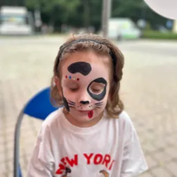 Dziewczynka z pomalowaną twarzą w motywie pieska, z zamkniętymi oczami i wystawionym językiem, siedzi na krześle, w tle rozmyty krajobraz.