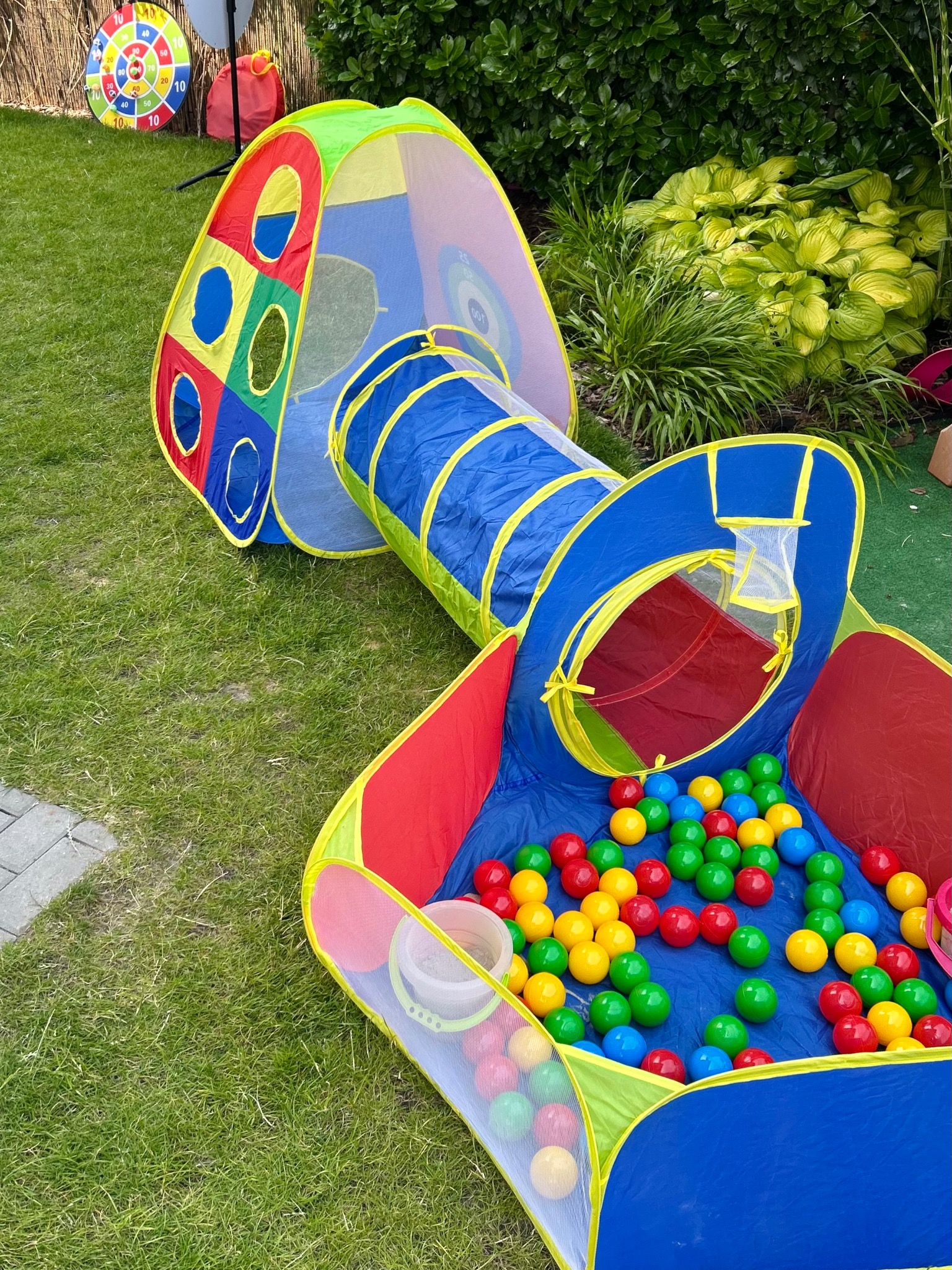Kolorowy tunel z basenem pełnym piłeczek na trawie, obok tarcza do rzutek. Atrakcja dla dzieci na imprezie plenerowej, animator dla dzieci.