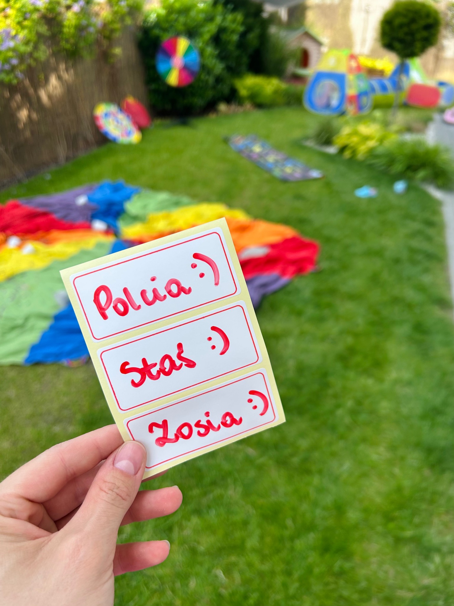 Nalepki z imionami 'Polcia', 'Staś' i 'Zosia' w dłoni na tle kolorowej chusty animacyjnej i placu zabaw na trawie. Przygotowania do zabawy z dziećmi.