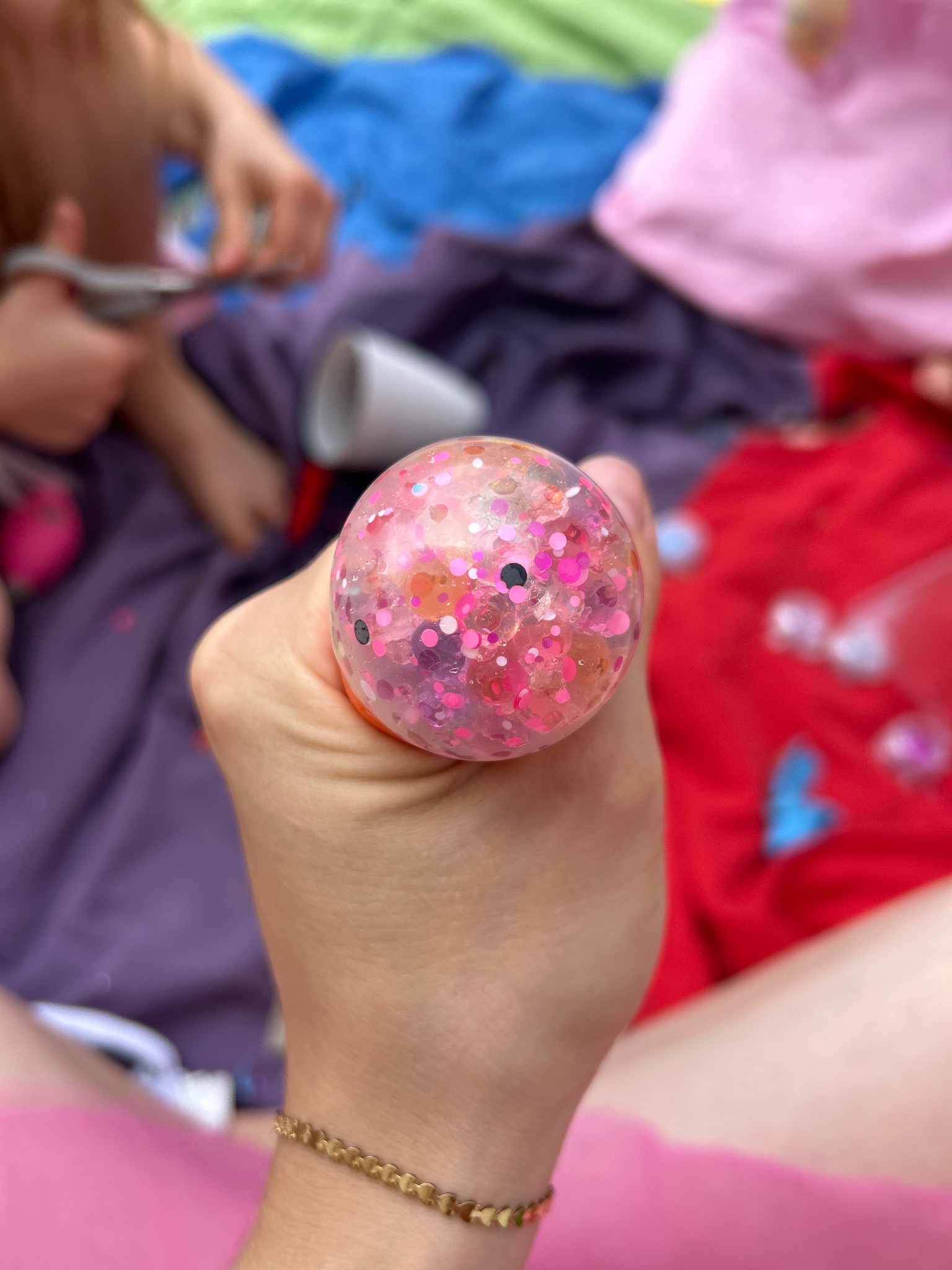 Dłoń trzymająca brokatową kulę na tle kolorowych materiałów i bawiących się dzieci, co sugeruje animację czasu wolnego.