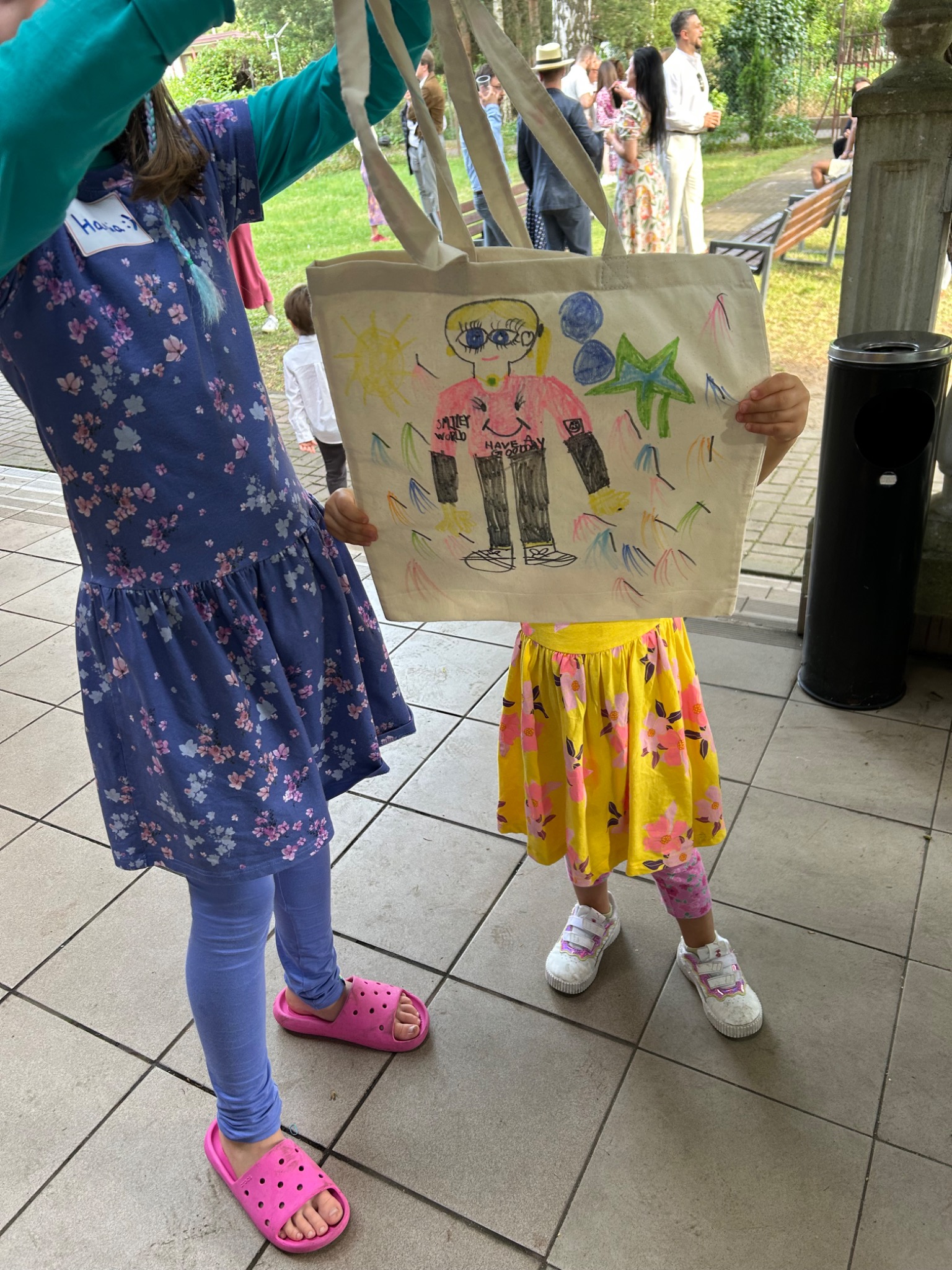 Dwie dziewczynki prezentują torbę z rysunkiem postaci z napisem 'Smiley World' i 'Have a good day', wykonanym prawdopodobnie podczas animacji dla dzieci.