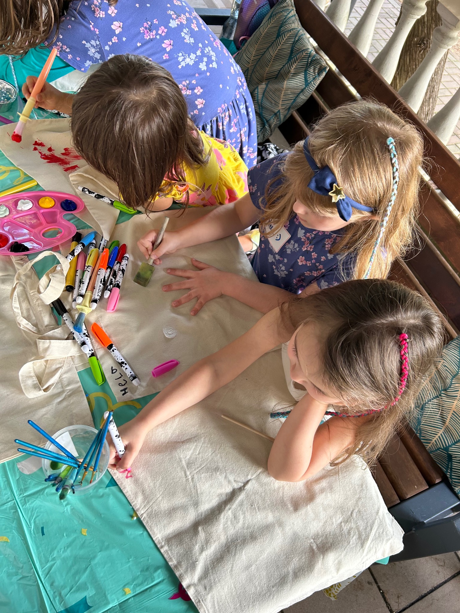 Dzieci malują farbami na płóciennych torbach, siedząc przy stole. Widoczne pędzle, markery i paleta kolorów. Kreatywne zajęcie dla dzieci podczas imprezy.