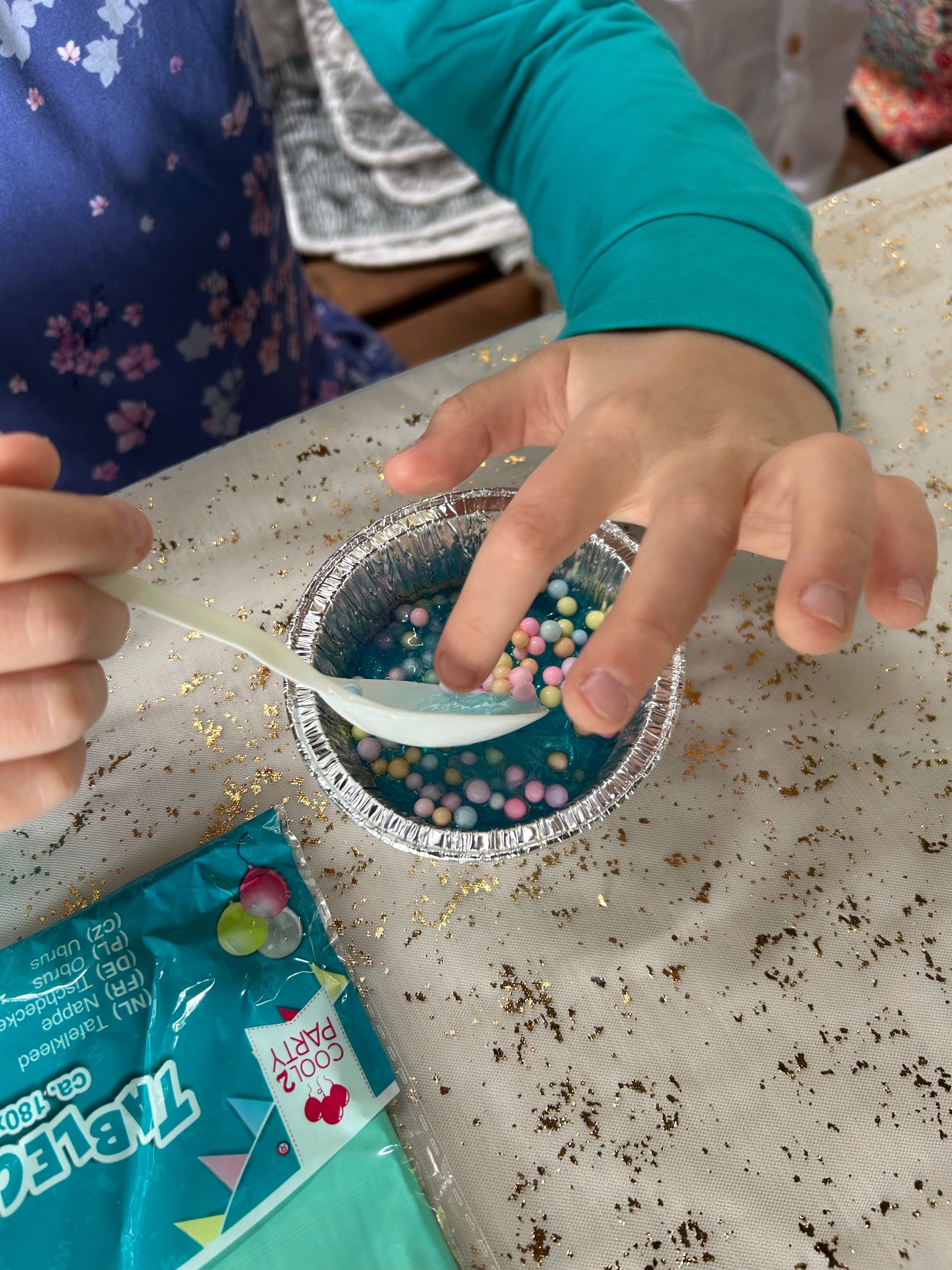 Dziecko miesza łyżką kolorowe kulki w niebieskim żelu w aluminiowym naczyniu na stole z dekoracją, obok leży opakowanie serwetek. Kreatywne zajęcie dla dzieci.