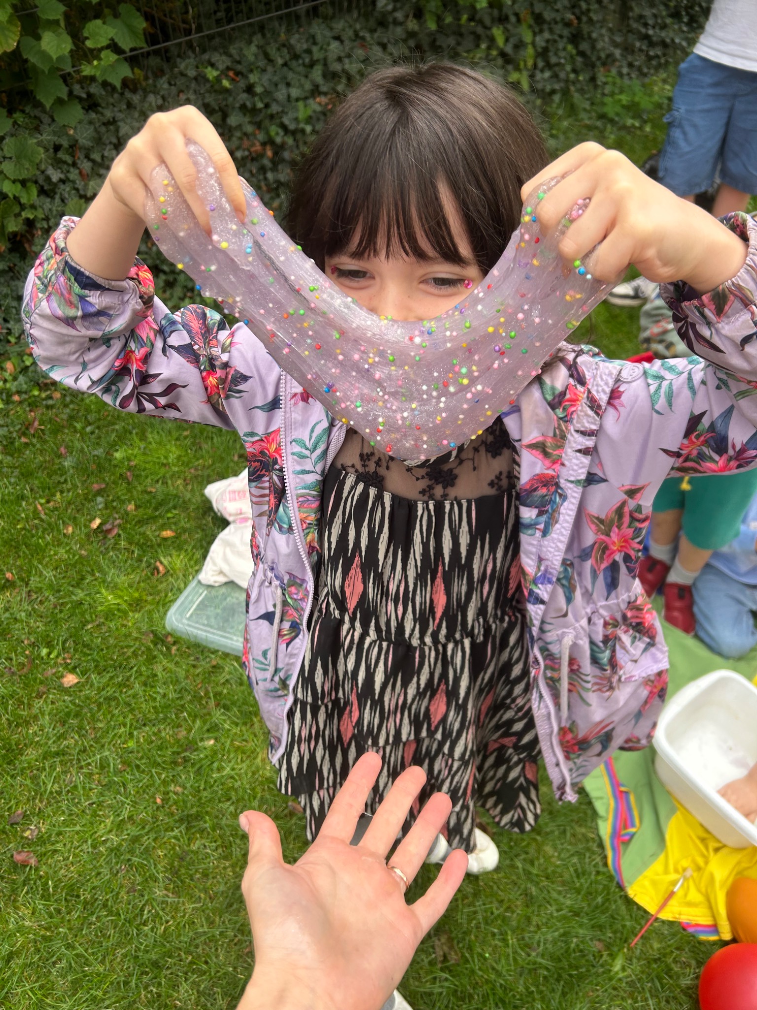 Dziewczynka z brokatowym slime na trawie, zabawa animacyjna na świeżym powietrzu z kolorowymi koralikami, kreatywne warsztaty dla dzieci, zbliżenie na slime.