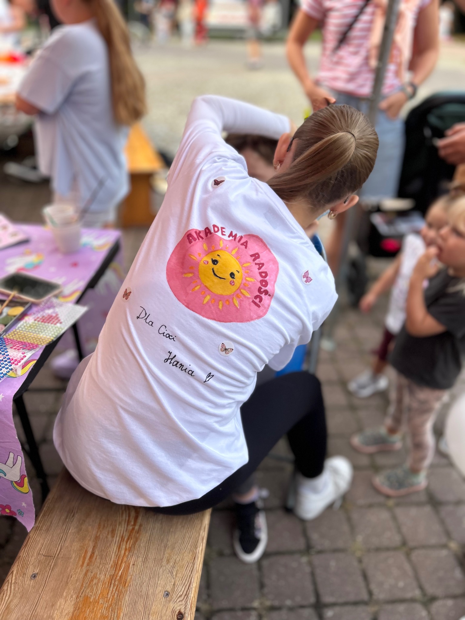 Kobieta w koszulce z logo 'Akademia Radości' w Konstancin-Jeziorna, siedzi na ławce. W tle dzieci i stolik z akcesoriami do animacji.