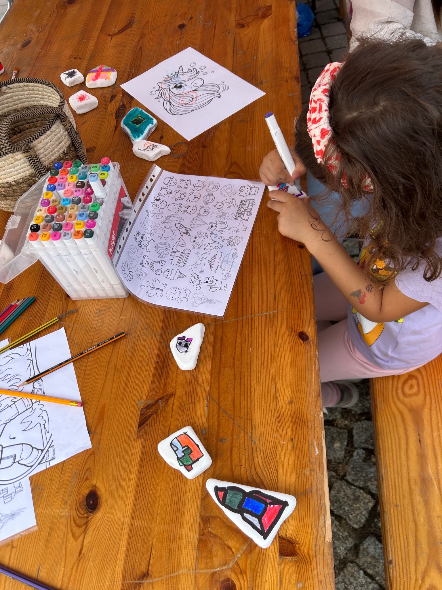 Dziewczynka z opaską maluje kolorowankę przy drewnianym stole z flamastrami, kredkami i pomalowanymi kamieniami. Kreatywne zajęcia na imprezie dla dzieci.