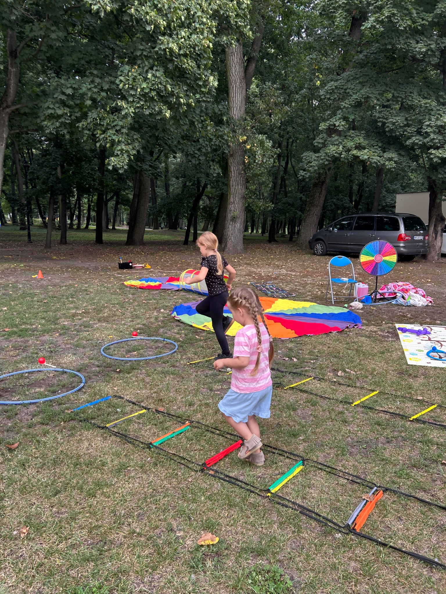 Dwie dziewczynki bawią się na trawie podczas animacji na świeżym powietrzu. Widoczne kolorowe rekwizyty, hula-hop i tor przeszkód. W tle drzewa i samochód.