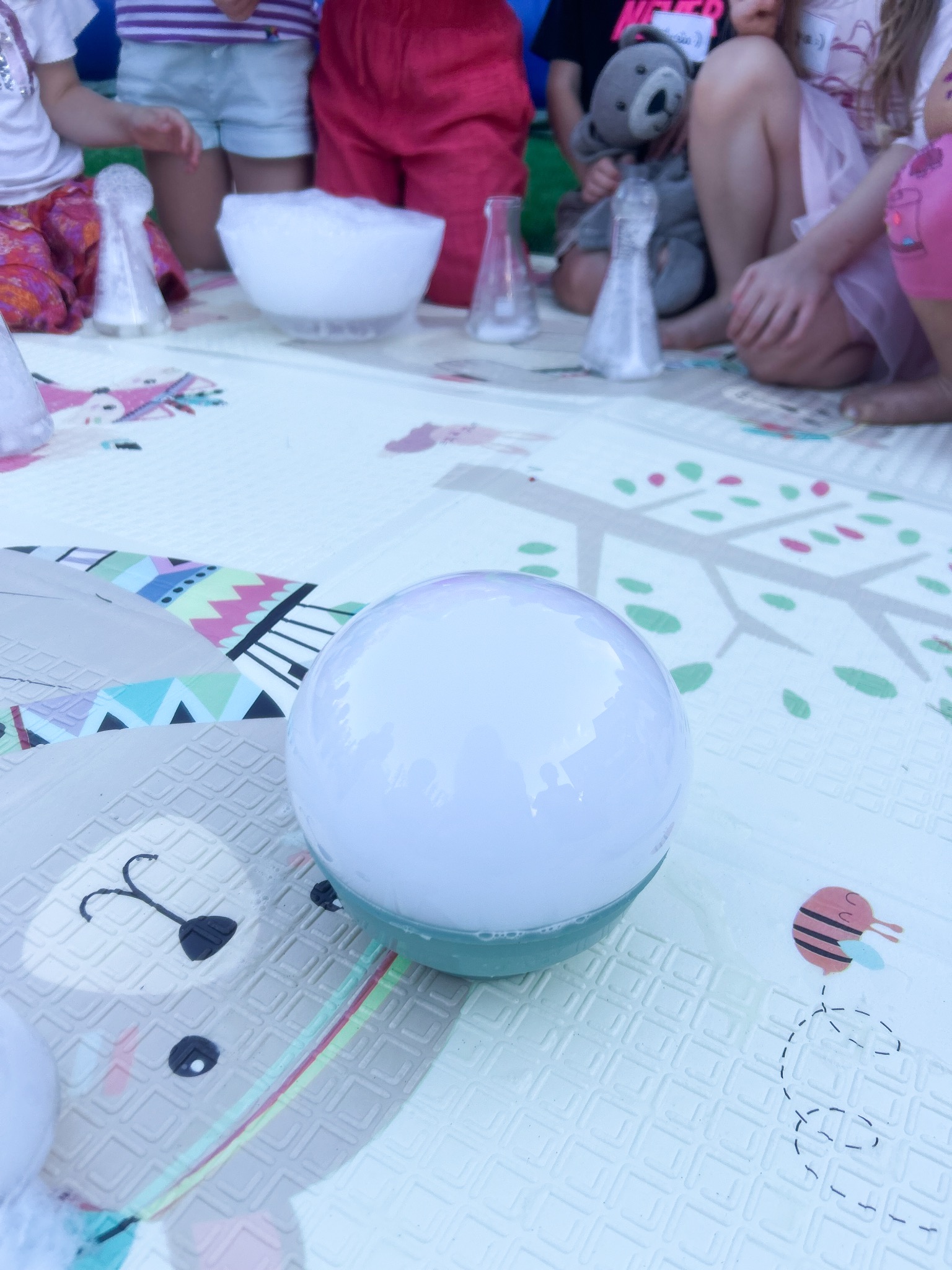 Dzieci obserwują eksperyment z bańkami mydlanymi na macie edukacyjnej. W tle kolby i miska z pianą. Zabawa z animatorem na imprezie dla dzieci.