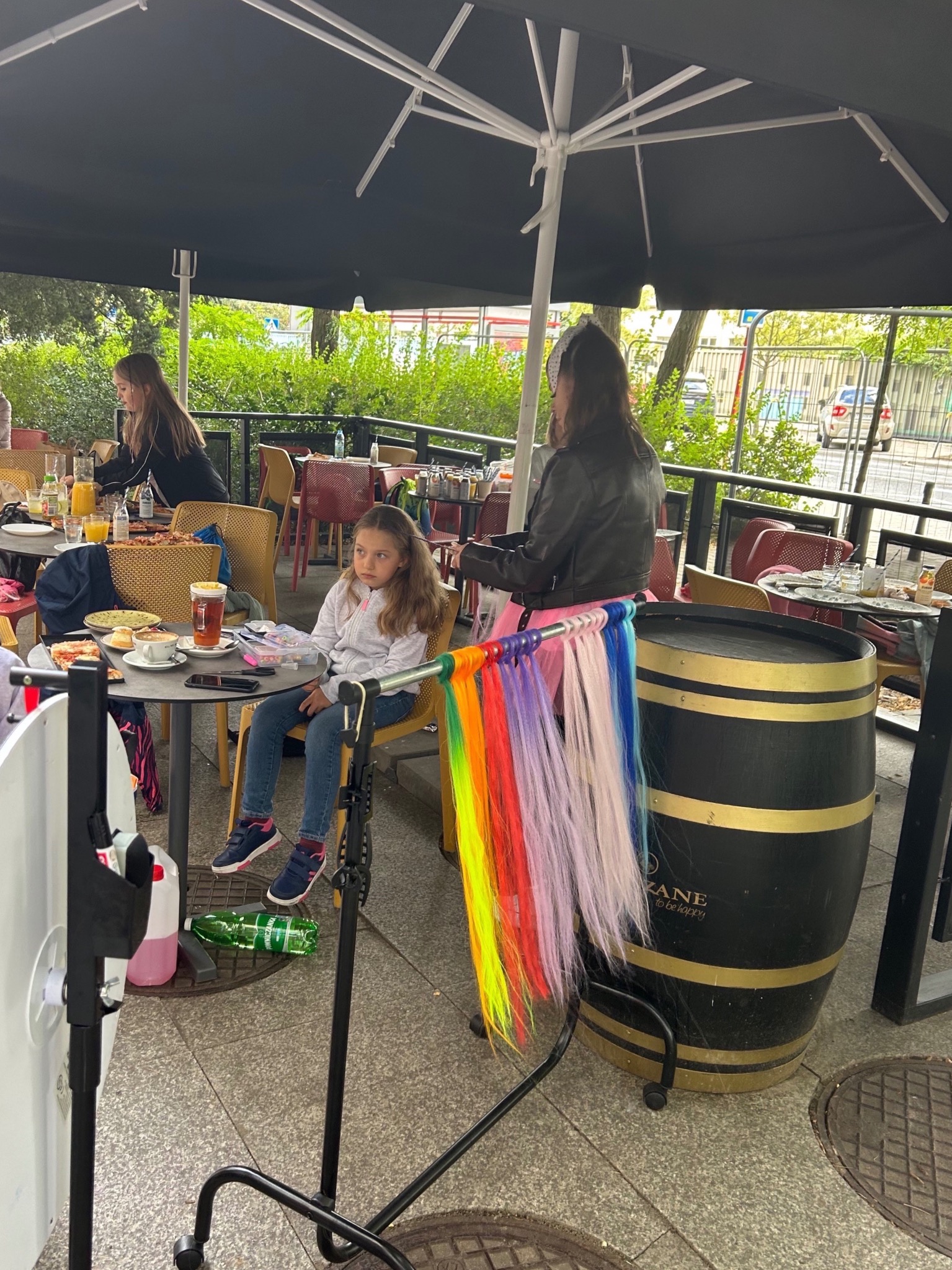 Dziewczynka siedzi przy stole z tęczowymi włosami doczepianymi obok, prawdopodobnie podczas imprezy plenerowej z animatorem.