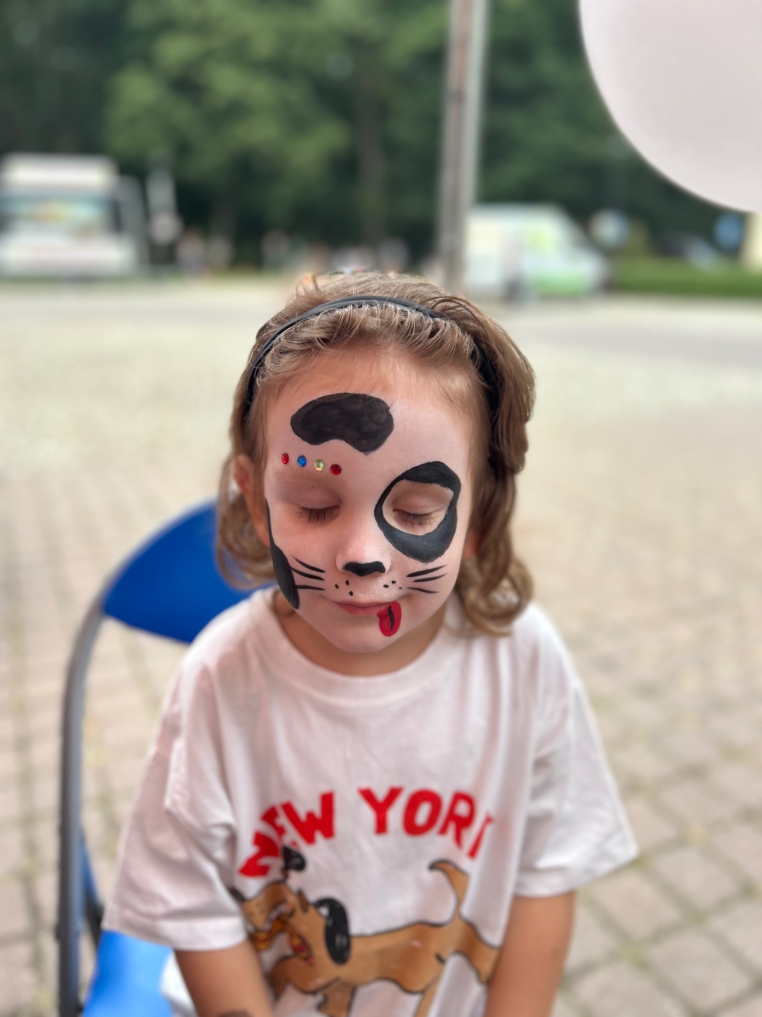 Dziewczynka z pomalowaną twarzą w motywie pieska, z zamkniętymi oczami i wystawionym językiem, siedzi na krześle, w tle rozmyty krajobraz.