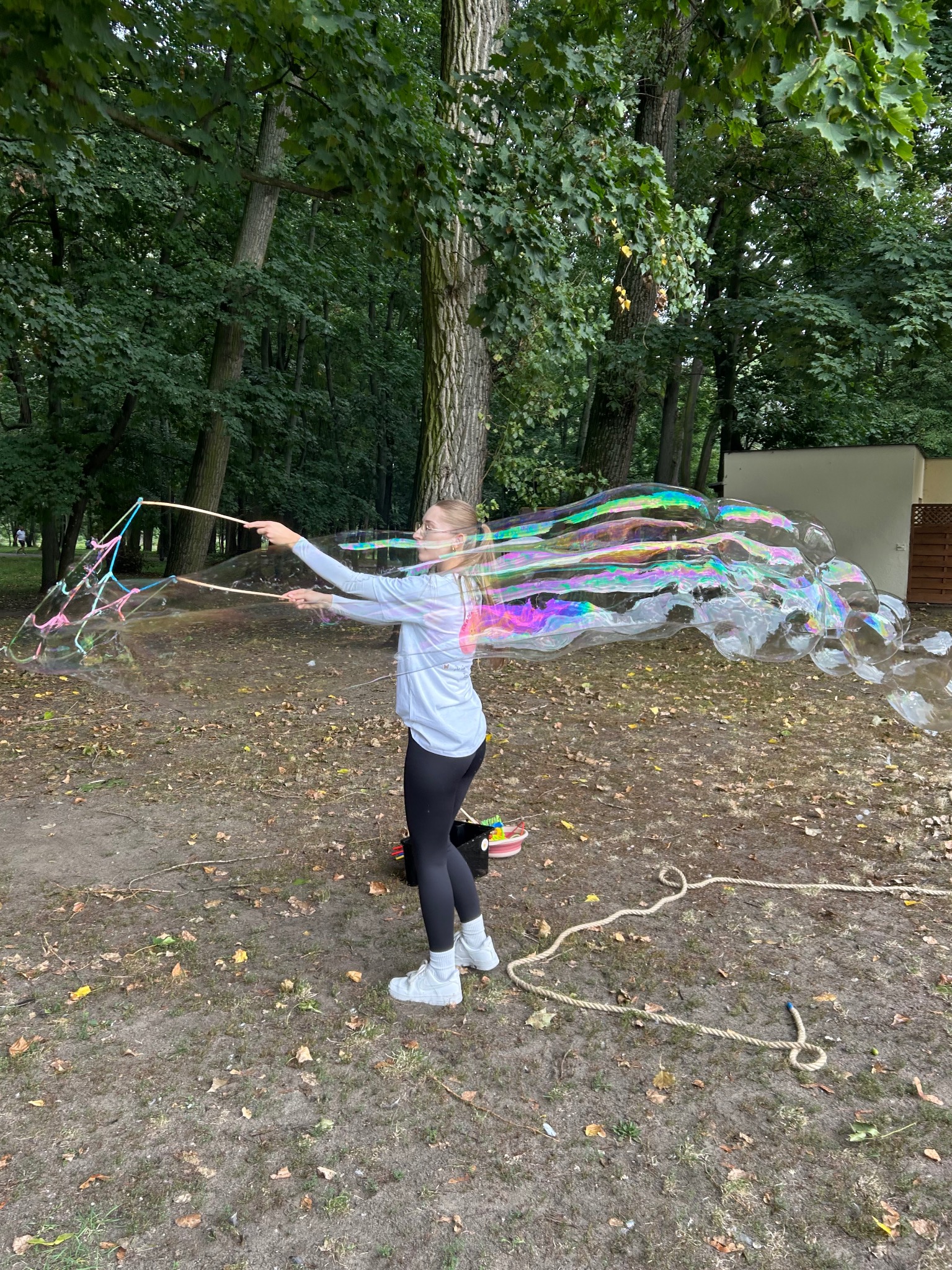 Kobieta tworzy gigantyczne bańki mydlane w parku, używając specjalnych sznurków i kijków. Widoczne tęczowe refleksy na bańkach i leśne tło.