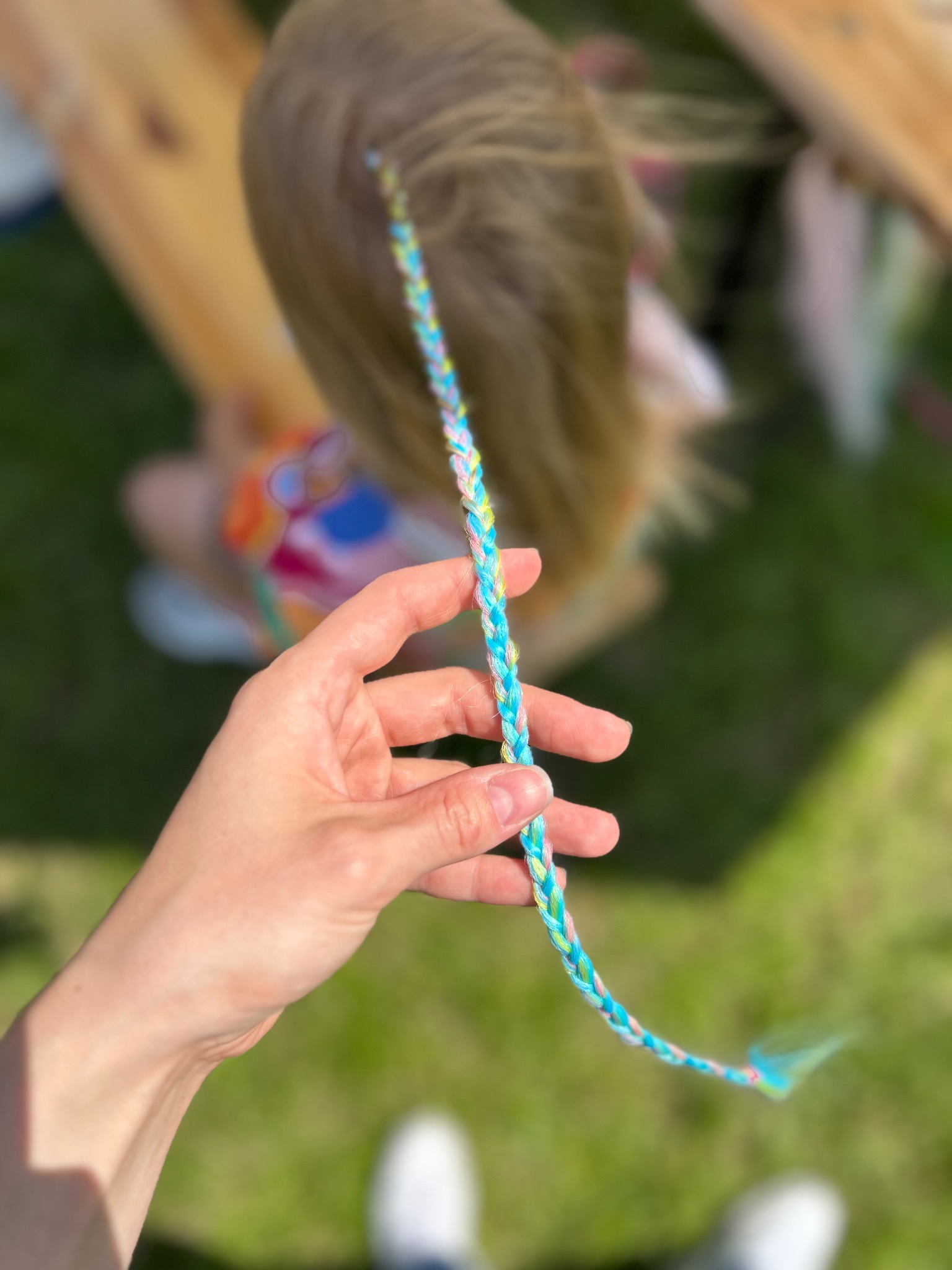 Kolorowa warkoczykowa ozdoba we włosach dziecka, trzymana w dłoni na tle zielonej trawy. Usługa stylizacji włosów dla dzieci na imprezie.