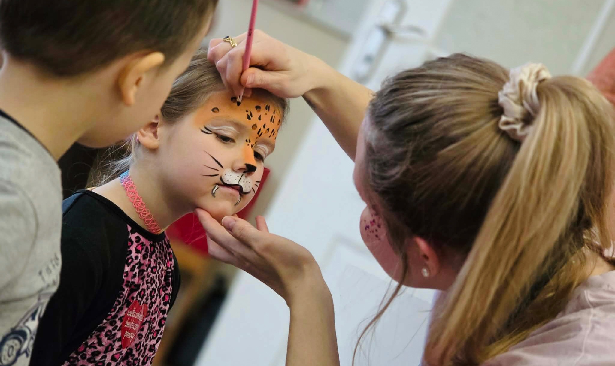 Malowanie twarzy dziewczynki na motyw zwierzęcy (gepard) przez animatora podczas imprezy. Widoczny pędzel i dłoń malującej. Dziewczynka ma różowy naszyjnik.