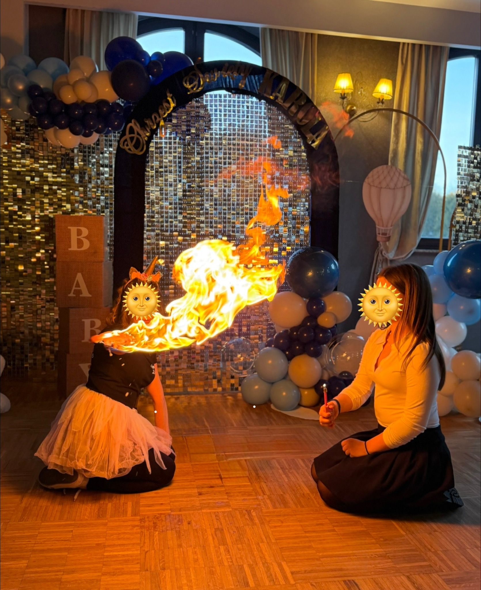 Dziewczynka w stroju dmucha ogniem na tle dekoracji urodzinowych z balonów i błyszczącej ścianki. Efektowny pokaz pirotechniczny na imprezie.