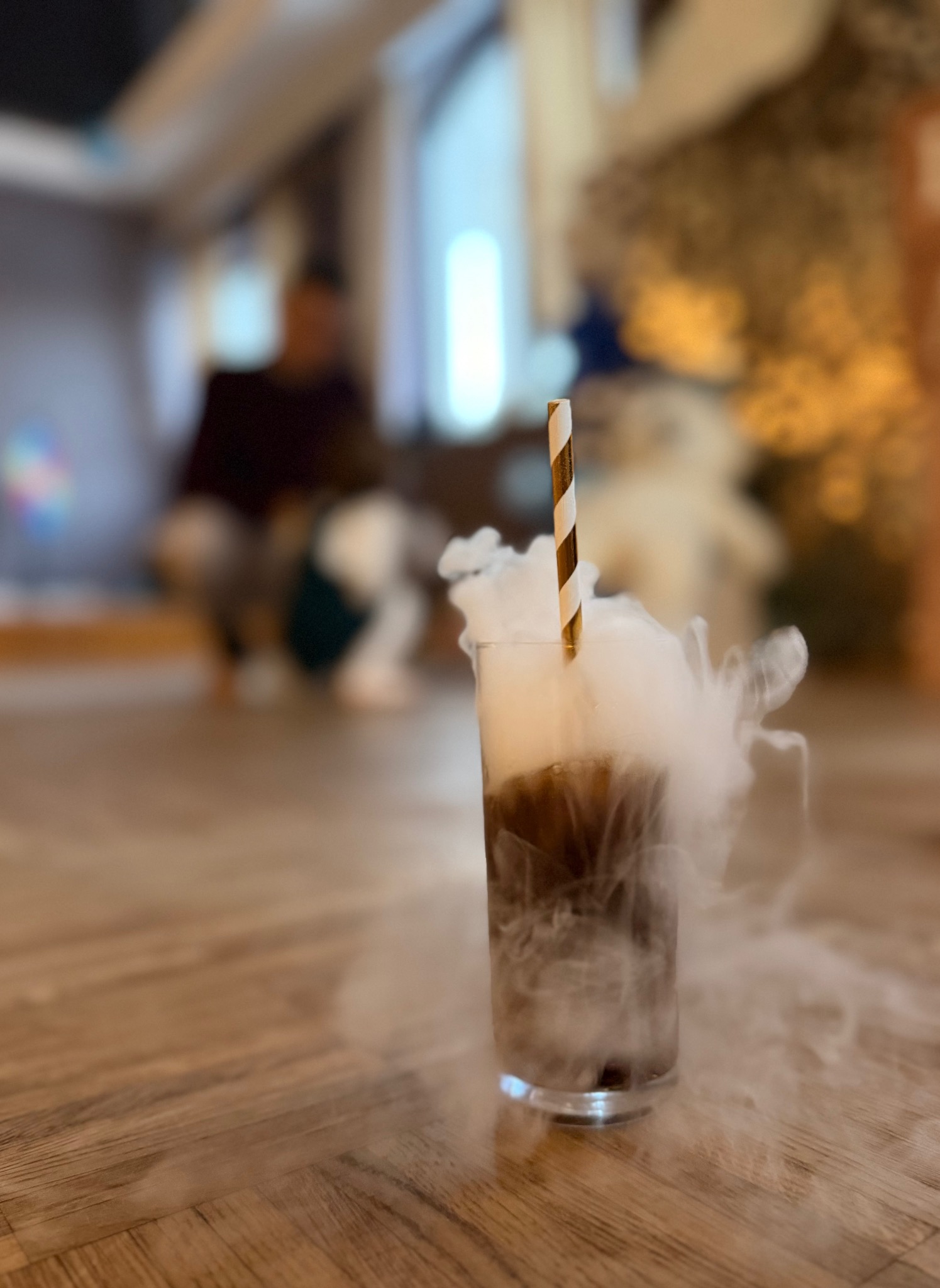 Efektowny napój z suchym lodem i złotą słomką na drewnianej podłodze, w tle postać w rozmyciu. Idealny na imprezę dla dzieci, tworząc magiczną atmosferę.