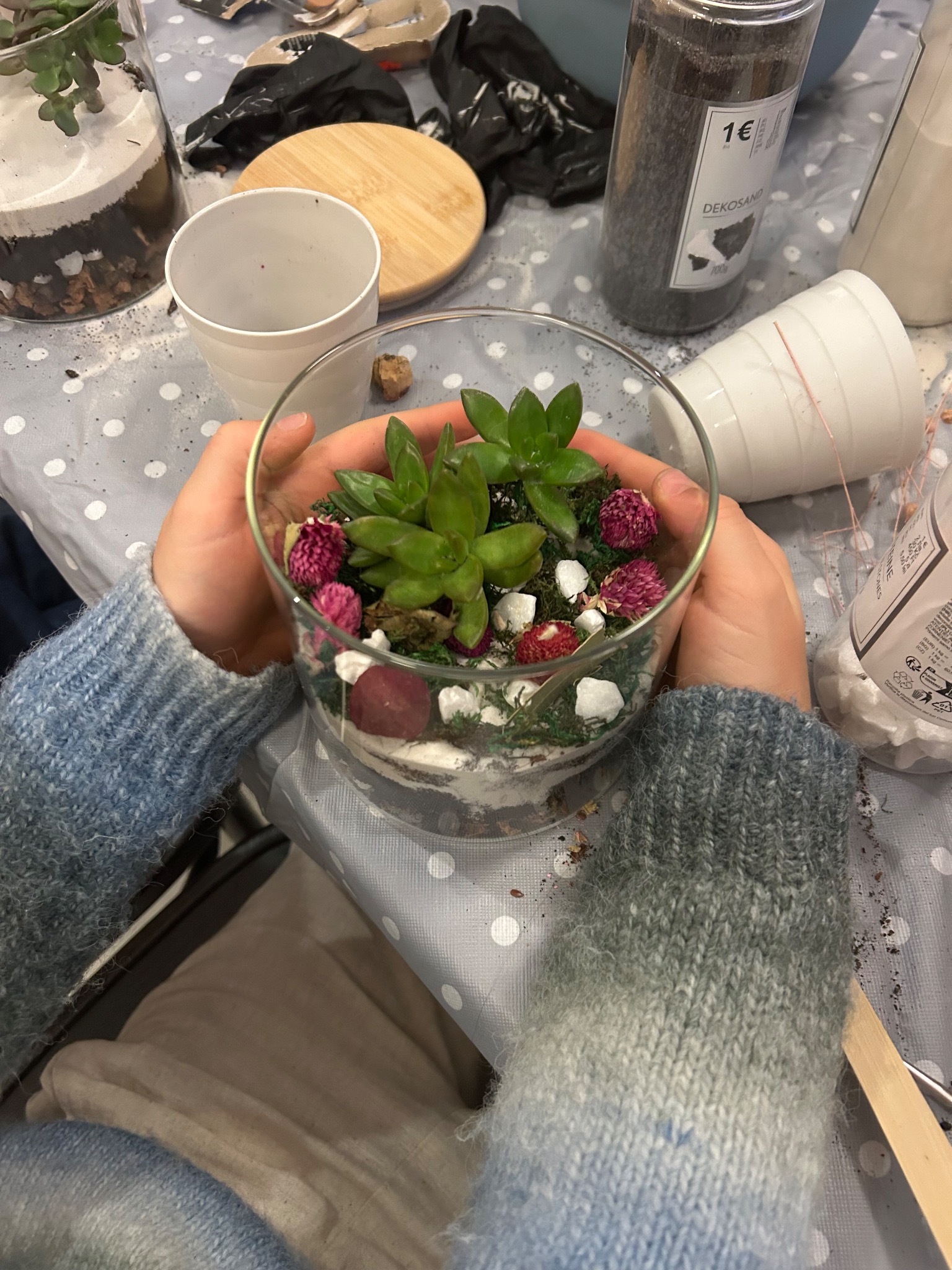 Szklane naczynie z sukulentami i dekoracjami w dłoniach na stole z akcesoriami do tworzenia mini-ogródka, warsztaty florystyczne dla dzieci.