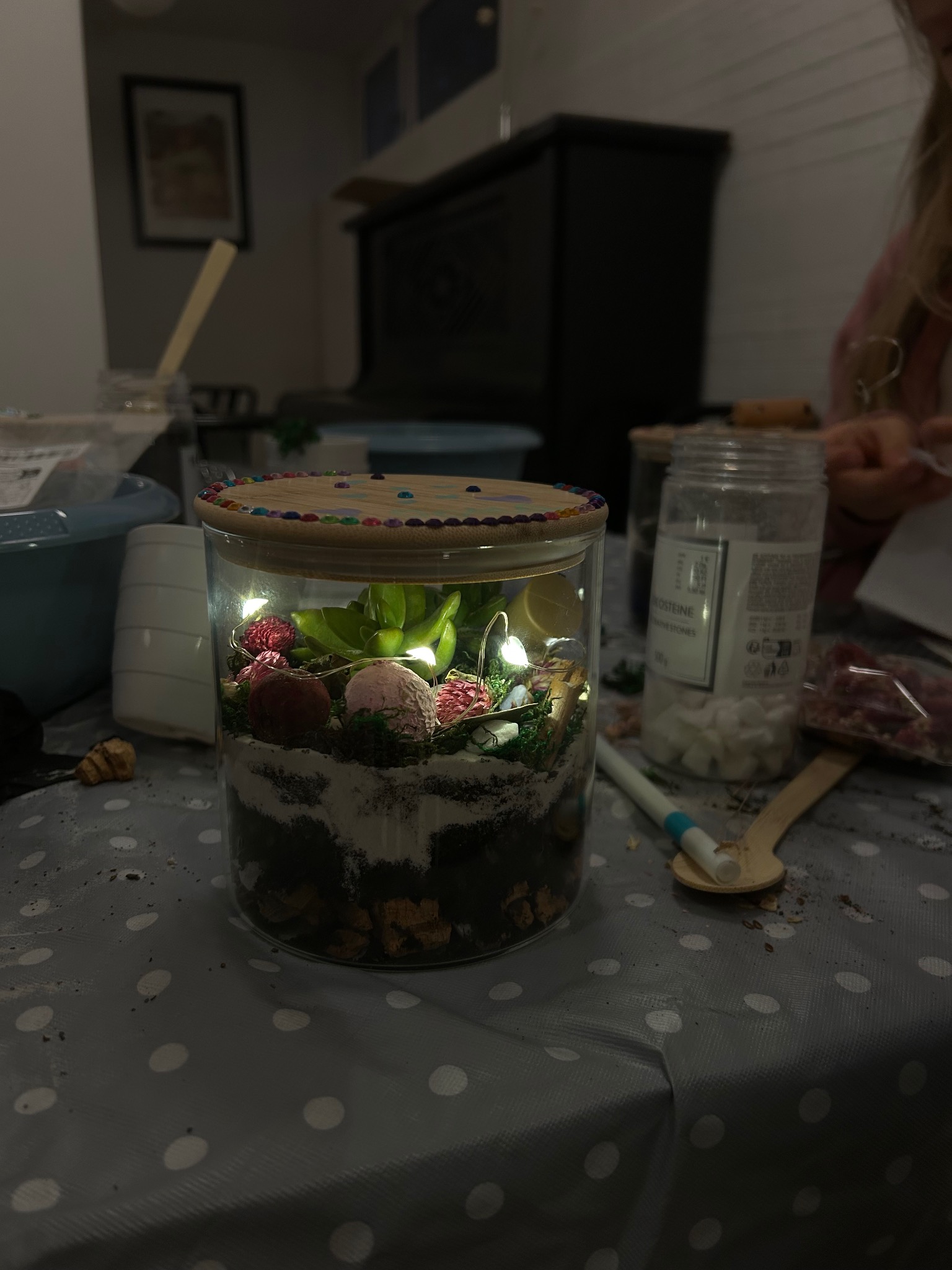 Szklany słoik z warstwami ziemi, roślin i dekoracji, oświetlony delikatnymi światełkami, na stole z akcesoriami do tworzenia mini-ogrodu.