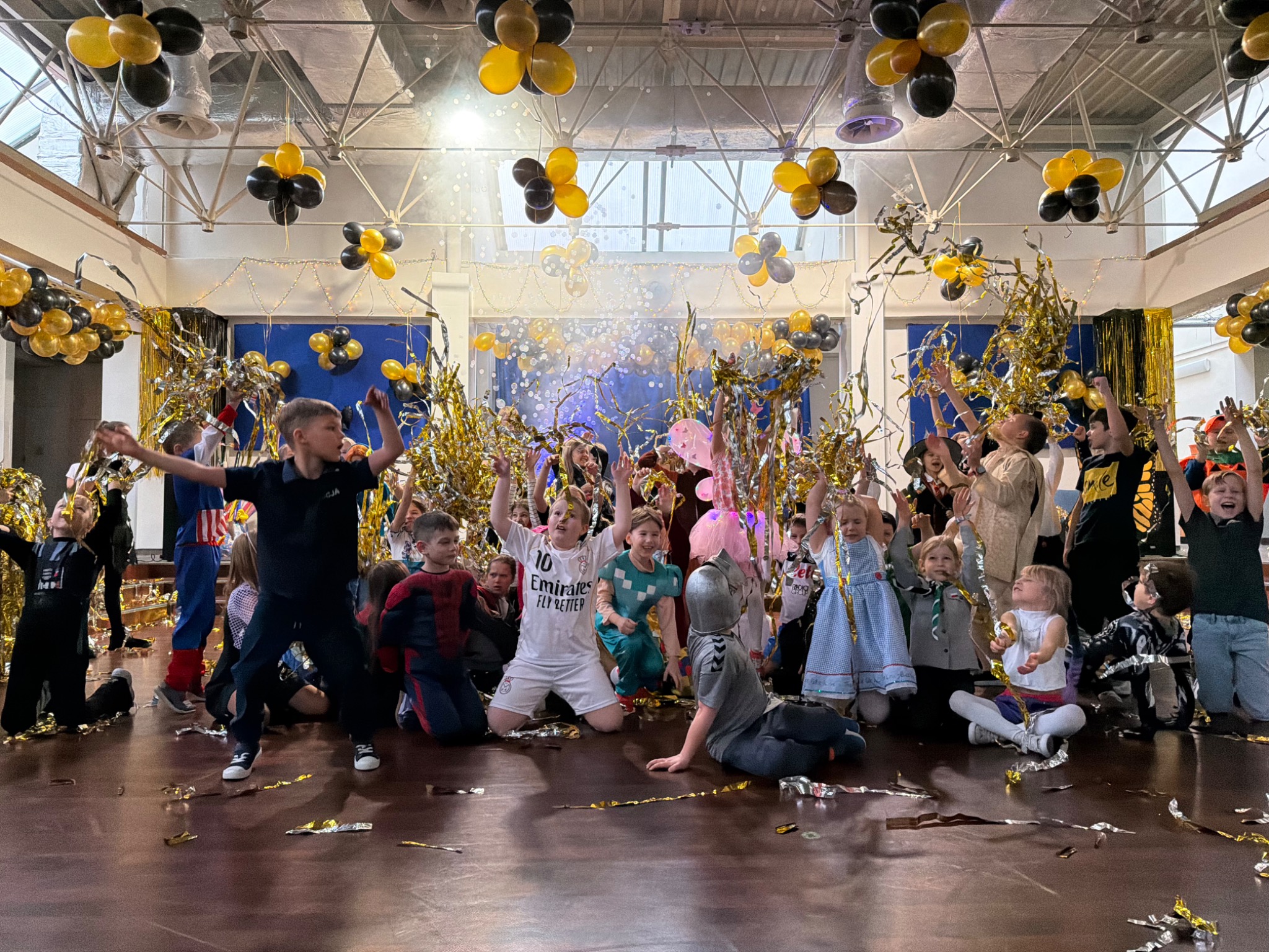 Uśmiechnięte dzieci w kostiumach bawią się złotym konfetti podczas imprezy z animatorem, w tle dekoracje z czarno-złotych balonów.