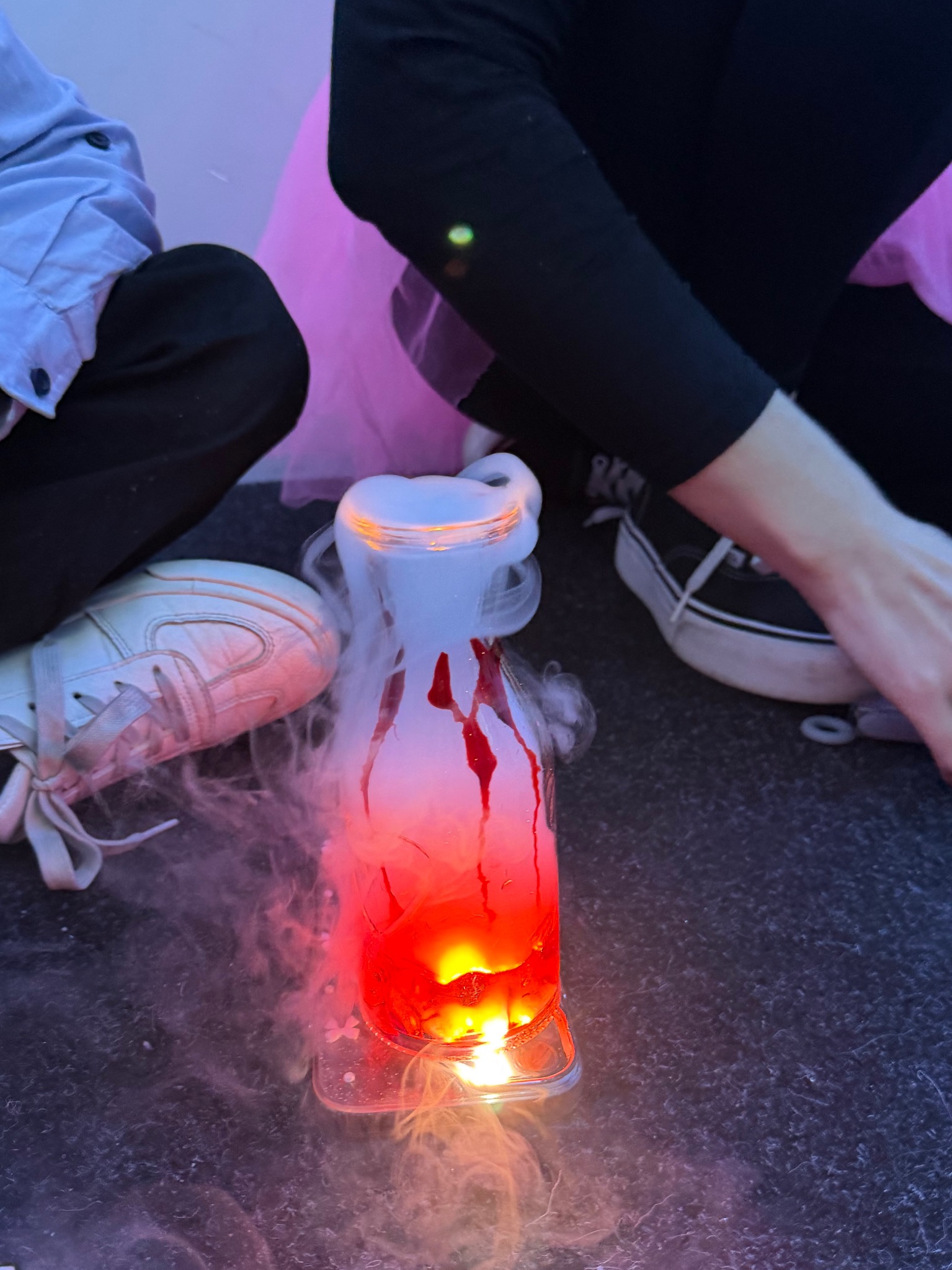 Eksperyment z dymiącą cieczą w szklanej butelce, podświetloną na czerwono, otoczoną przez dziecięce buty i fragment różowej tiulowej spódniczki.