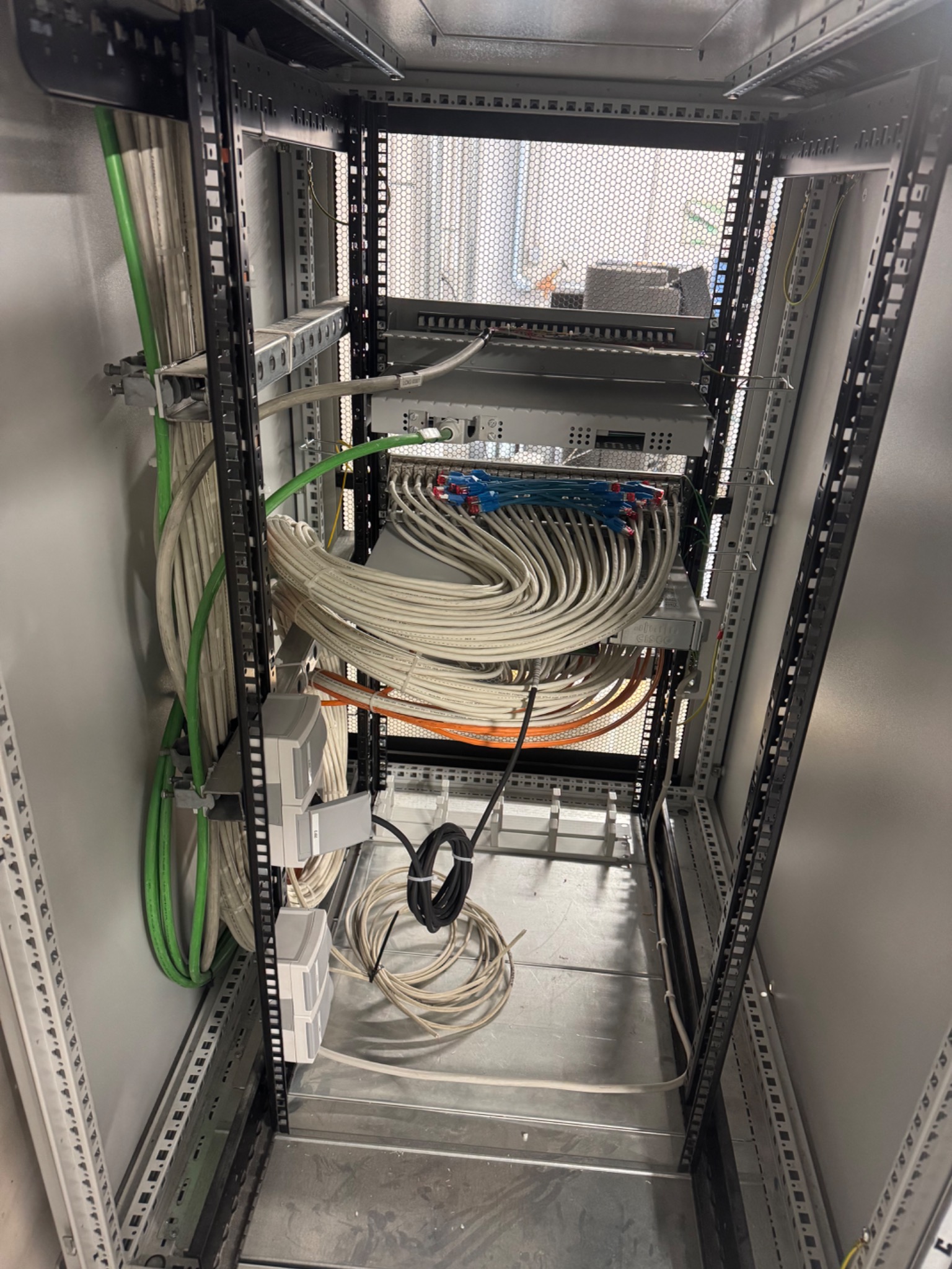 Wnętrze szafy serwerowej z uporządkowanymi kablami sieciowymi (białe, zielone, pomarańczowe) i urządzeniami Cisco, widok z dołu, szara obudowa.