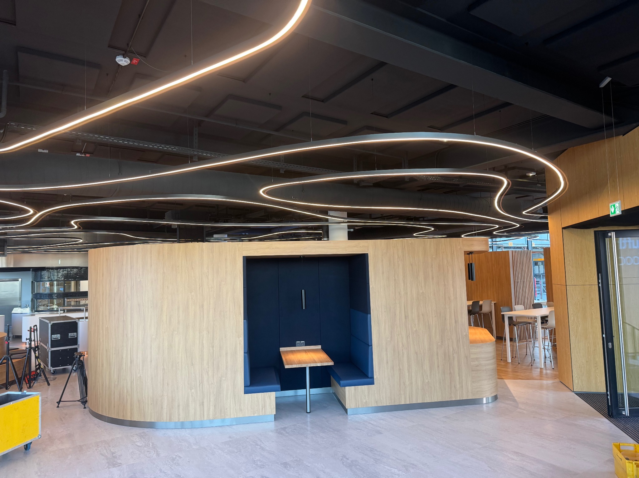 Nowoczesne oświetlenie LED w biurze: zakrzywione linie na ciemnym suficie, drewniane ścianki działowe z wnęką i niebieskimi siedziskami, jasna podłoga.