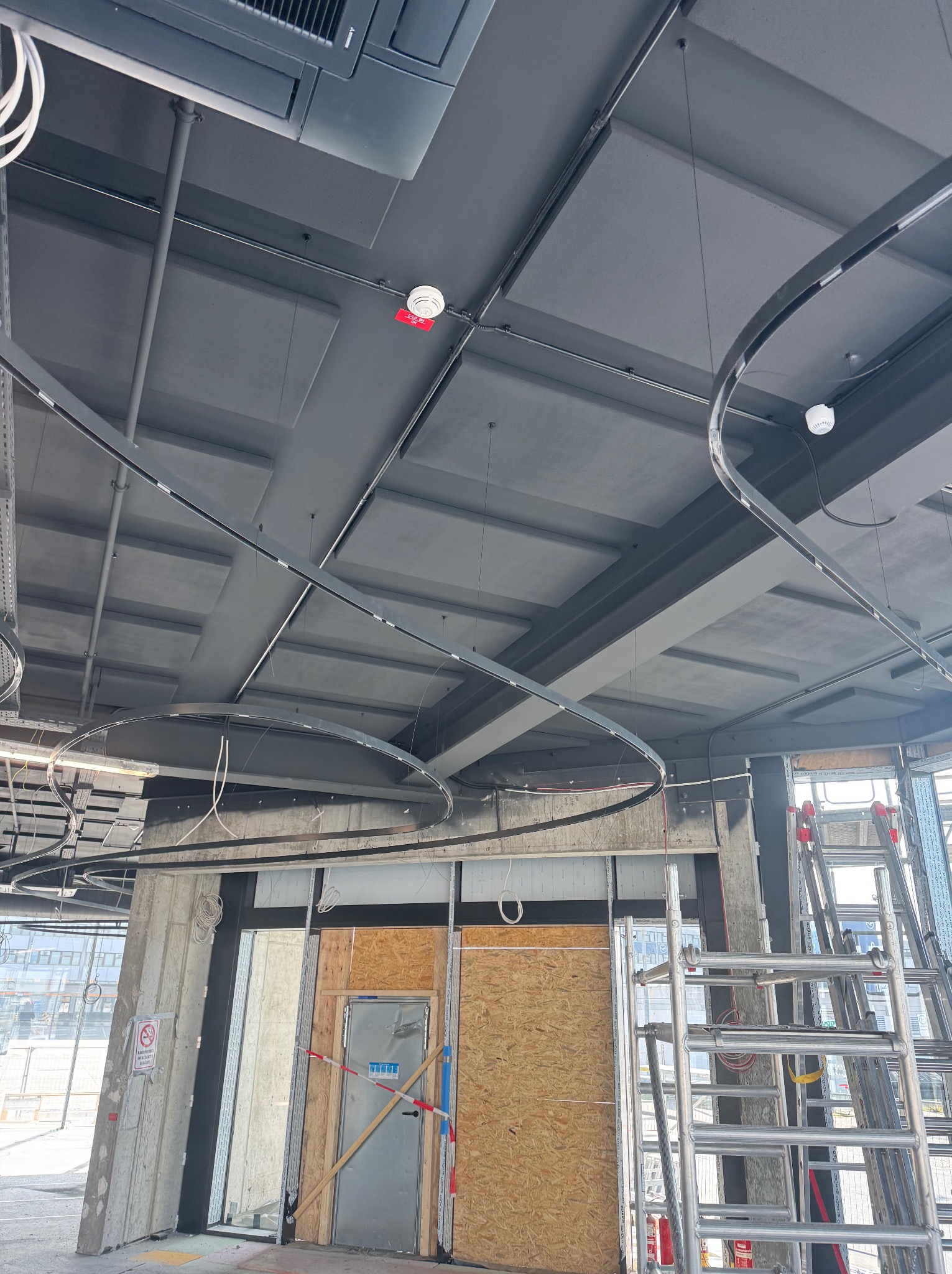 Szara sufitowa instalacja z elementami oświetlenia i przewodami w budynku w trakcie wykończenia, widoczne rusztowania i elementy konstrukcyjne.