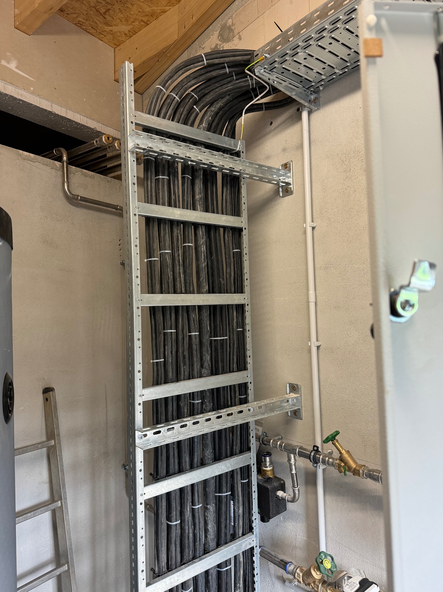 Rząd kabli w metalowej korycie przymocowanej do ściany, widoczne rury i elementy instalacji wodnej oraz drabina. Surowy stan wykończenia wnętrza.