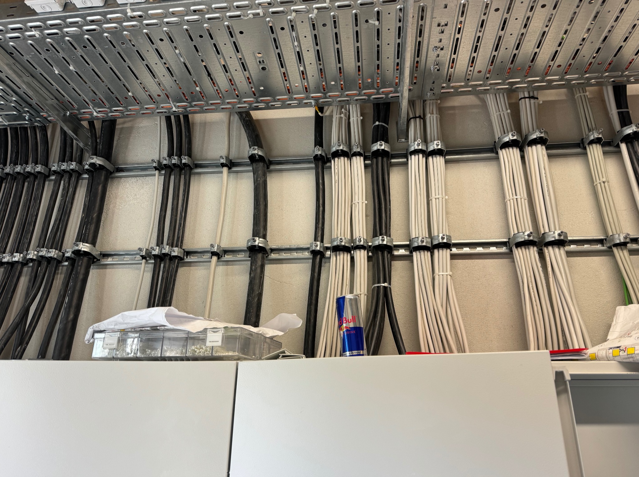 Równolegle ułożone kable elektryczne, czarne i białe, przymocowane do metalowej konstrukcji. Na szafce narzędzia i napój energetyczny.