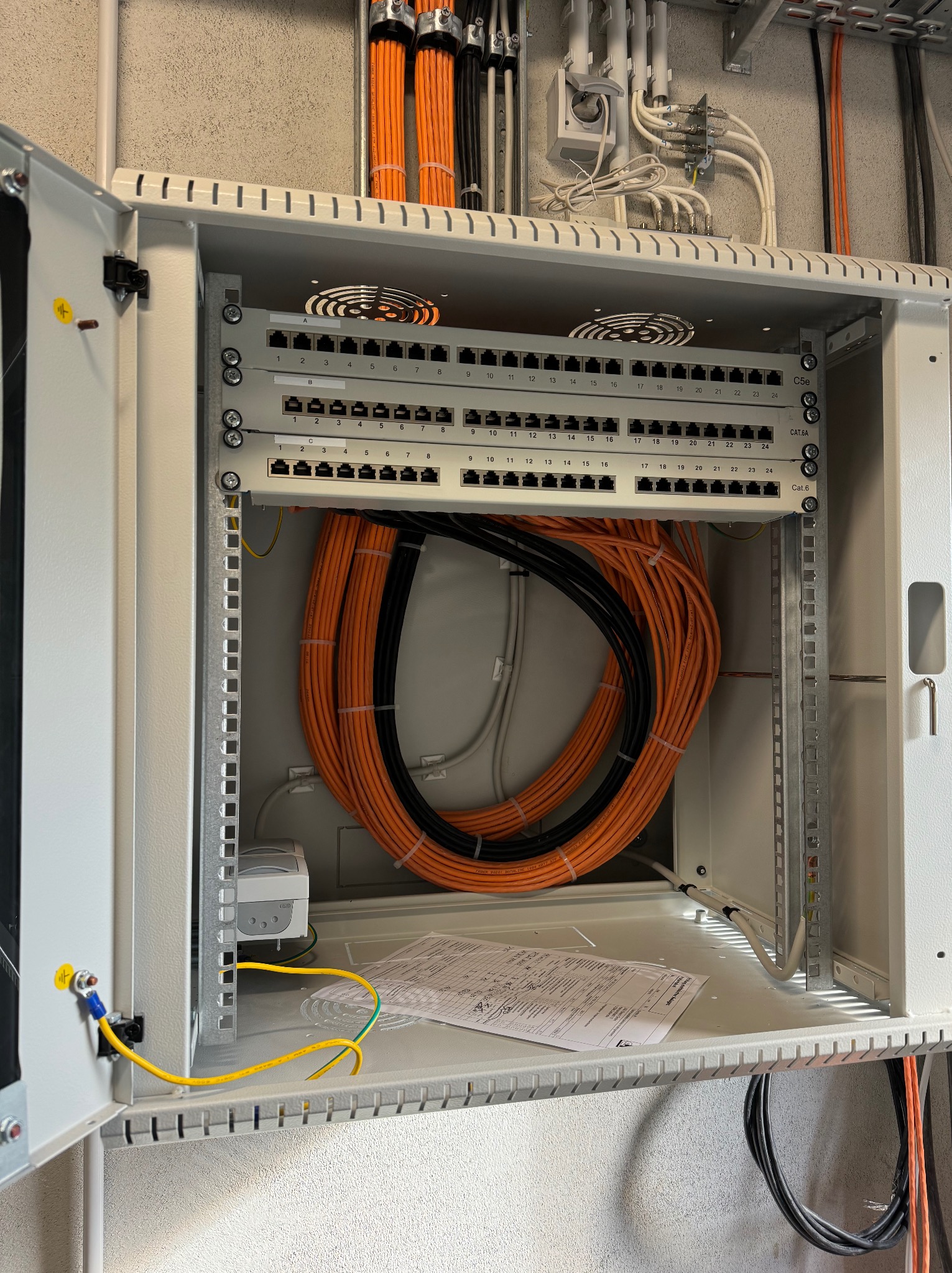 Otwarta szafa rack z panelami krosowniczymi Cat.6 i Cat.6A, okablowaniem strukturalnym (pomarańczowe i czarne kable) oraz uziemieniem. Widok na instalację.