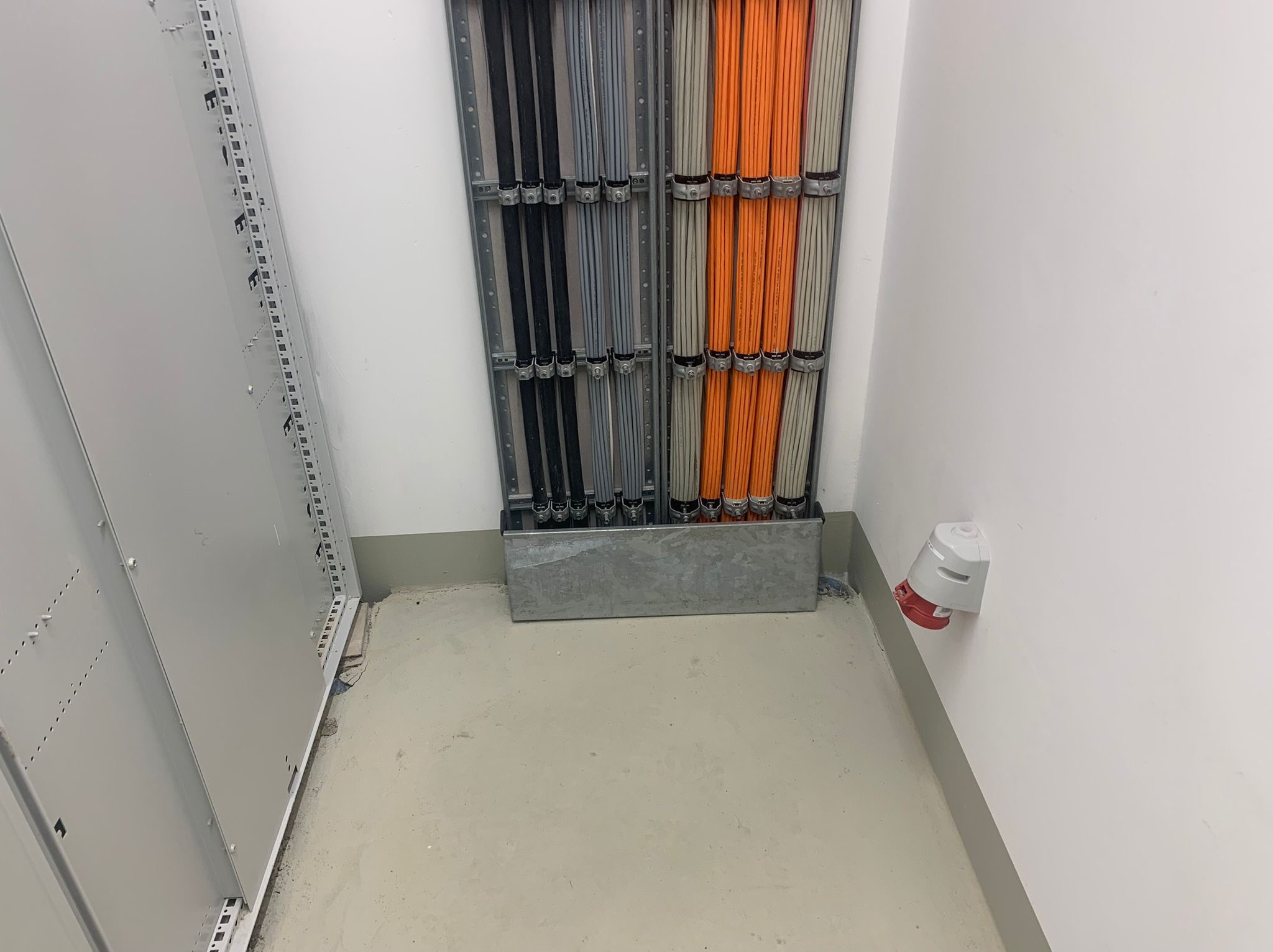 Uporządkowane kable elektryczne w korytach, czarne, szare i pomarańczowe, zamocowane do ściany w pomieszczeniu technicznym. Widoczna szafa rack i gniazdo elektryczne.