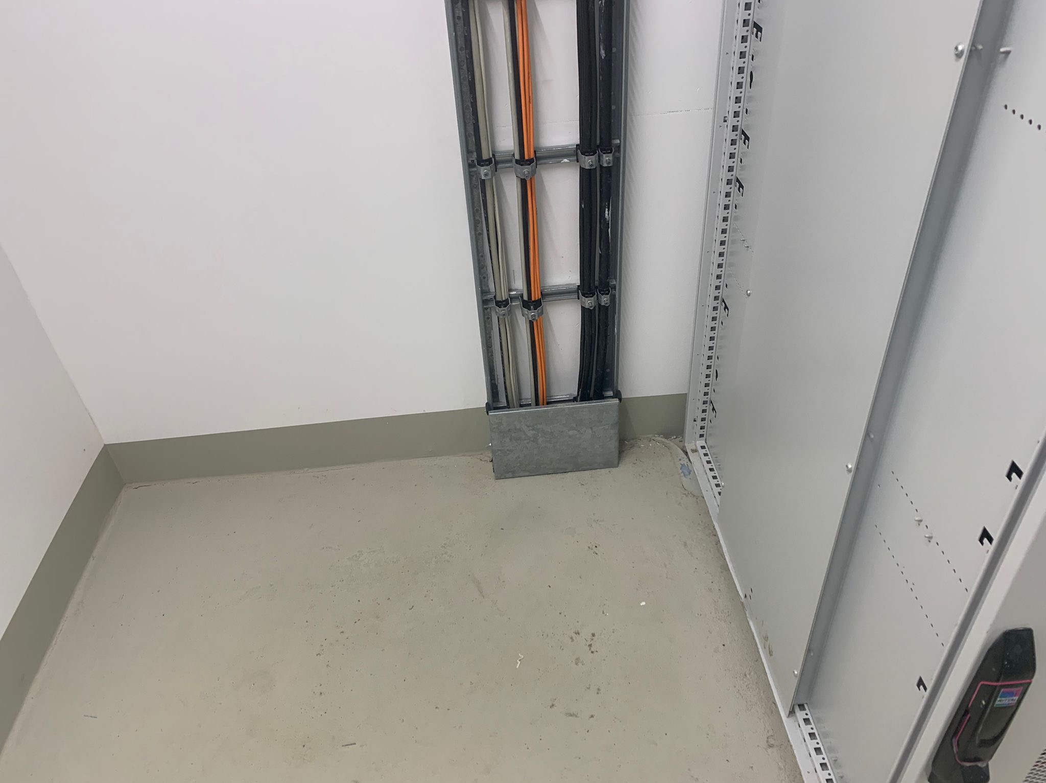 Kable elektryczne prowadzone w metalowej korycie na ścianie w pomieszczeniu technicznym. Widoczne przewody w kolorach: czarnym, pomarańczowym i srebrnym.