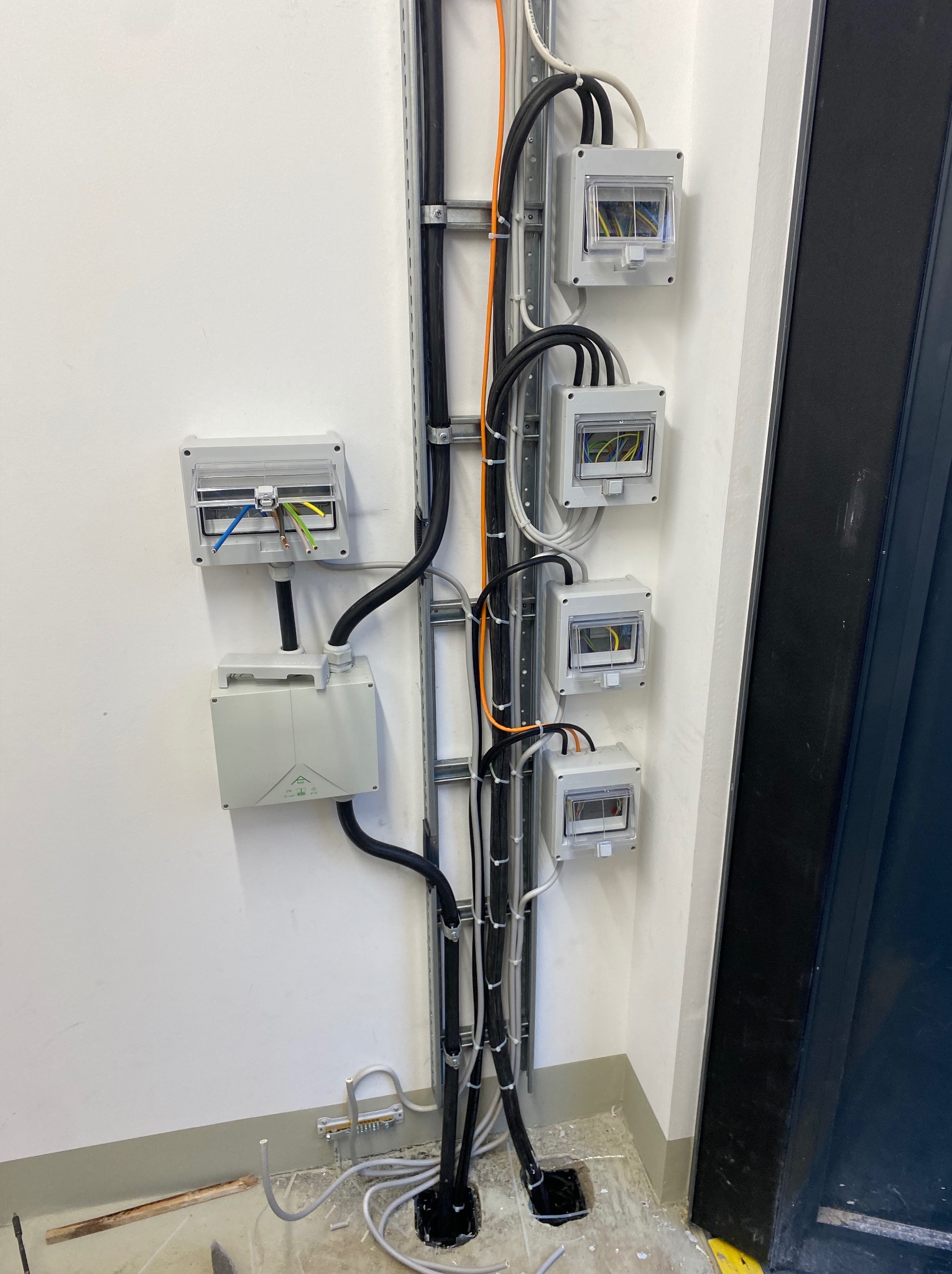 Szara metalowa listwa z przymocowanymi przewodami i puszkami elektrycznymi na białej ścianie, widoczne kable wewnątrz puszek.