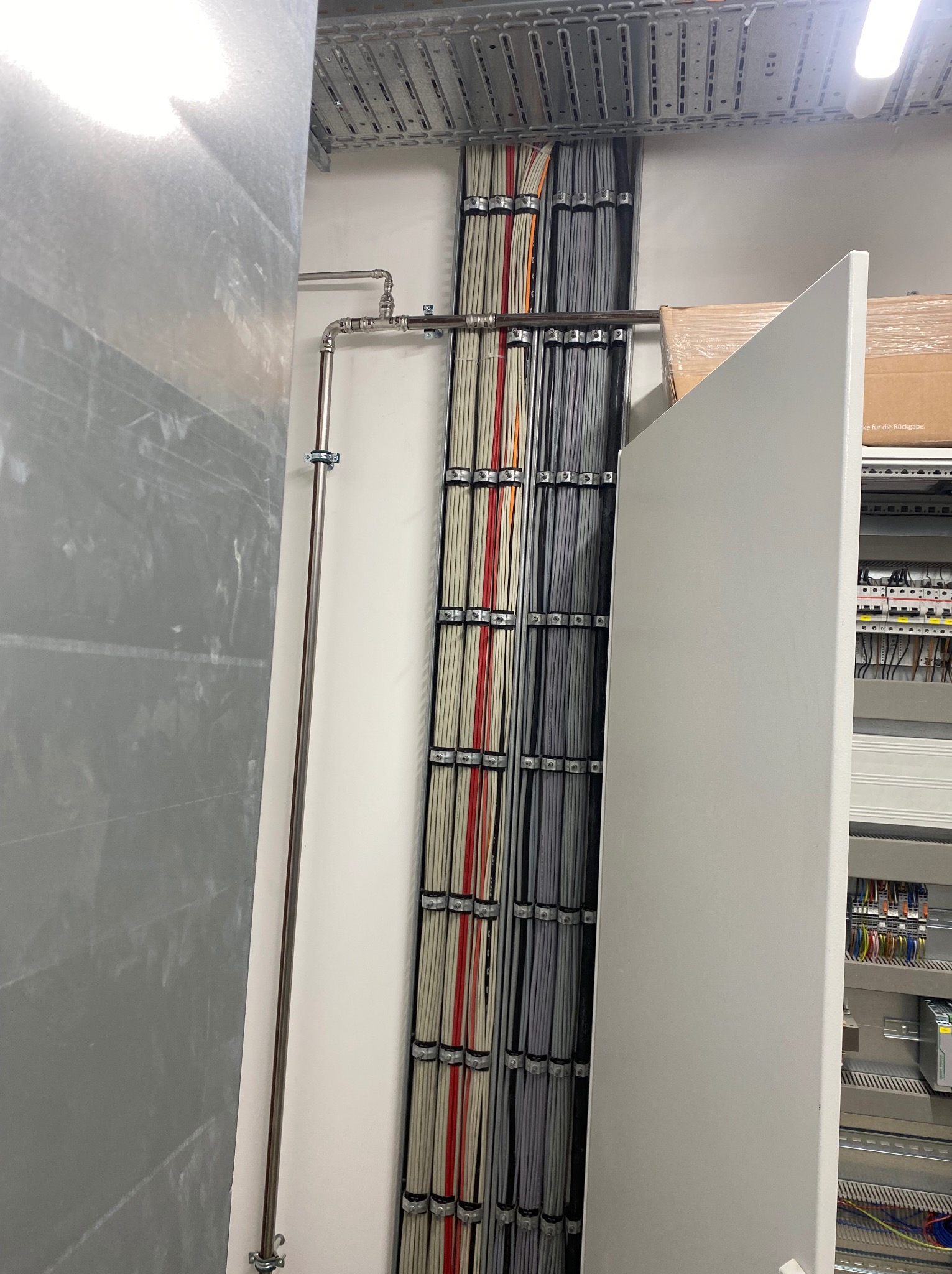 Rzędami ułożone kable elektryczne w korytkach, zabezpieczone uchwytami, obok metalowa rurka przymocowana do ściany. Widoczna szafa sterownicza z bezpiecznikami.