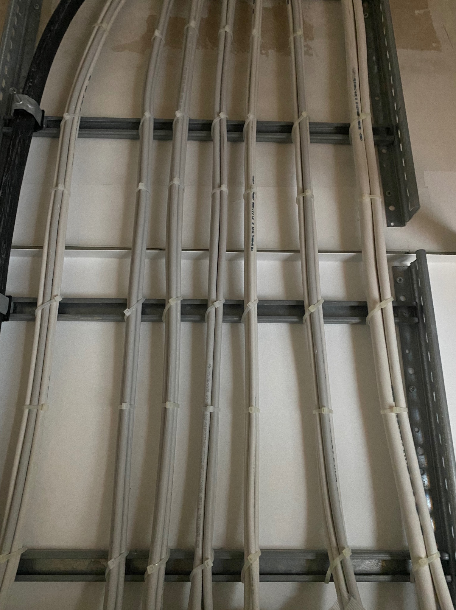 Ułożone równolegle przewody elektryczne, zabezpieczone opaskami, zamocowane do metalowej konstrukcji na jasnej ścianie. Widoczne fragmenty czarnego kabla.