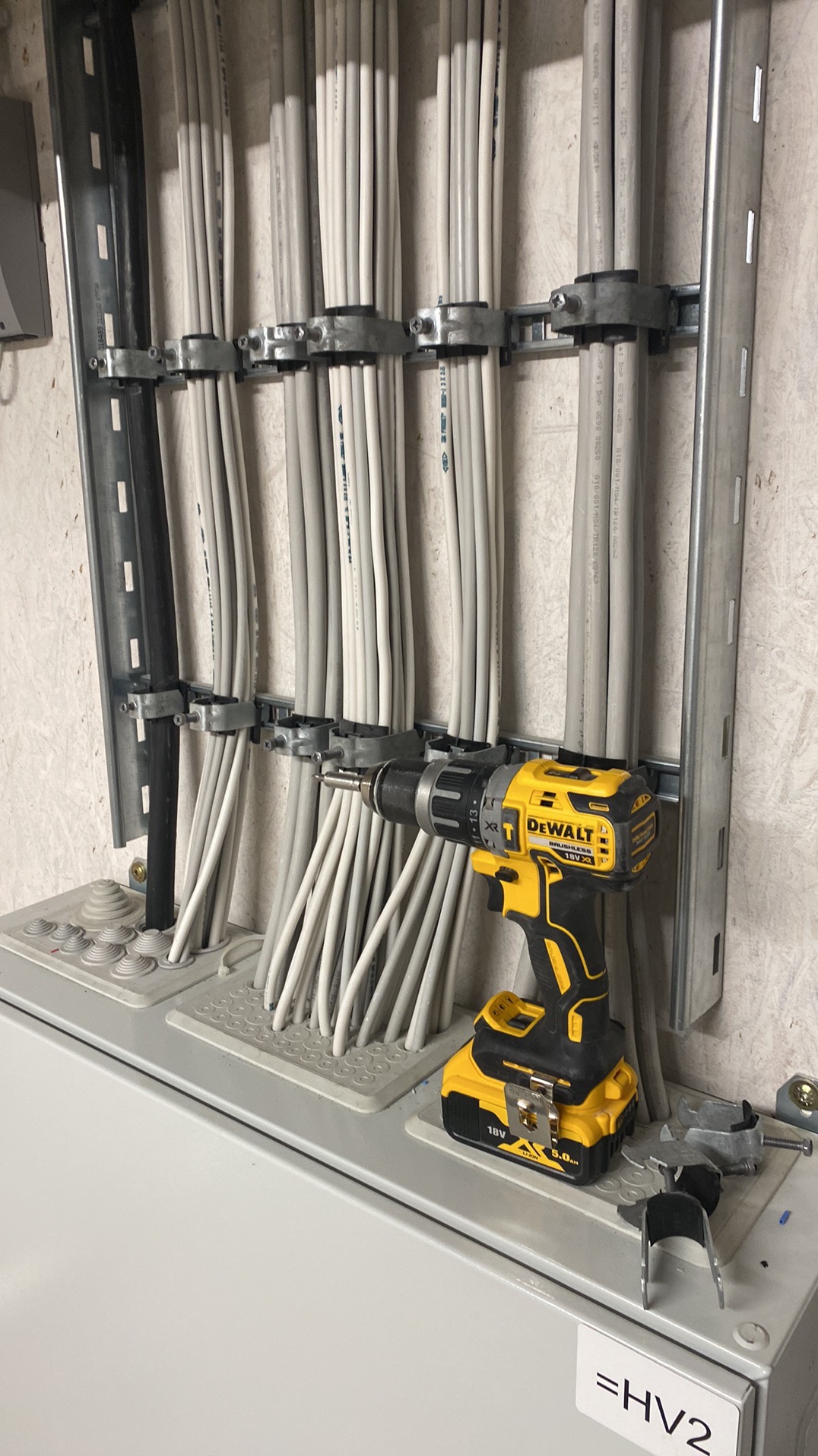 Szereg szarych kabli elektrycznych, przymocowanych do metalowej konstrukcji, z żółto-czarną wkrętarką DeWalt. Instalacja w trakcie prac.
