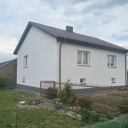 Biały dom z brązowym dachem po renowacji elewacji, widoczny zza czerwonego ogrodzenia, z tablicą miejscowości w tle i budynkiem gospodarczym z lewej.