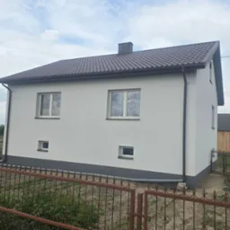 Dwukondygnacyjny dom z ocieploną, jasną elewacją i brązowym dachem z blachodachówki, widziany zza metalowego płotu. Widoczne okna i fragment posesji.