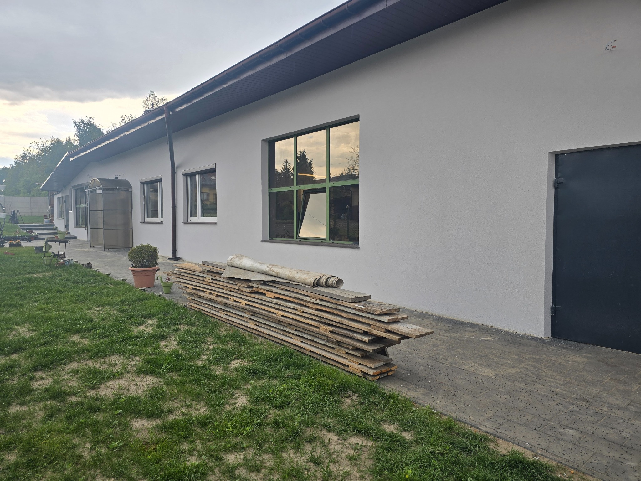 Biały budynek po renowacji elewacji, z oknami w zielonych ramach i stertą desek na chodniku. Widok na budynek z zewnątrz, trawnik i drzewa w tle.