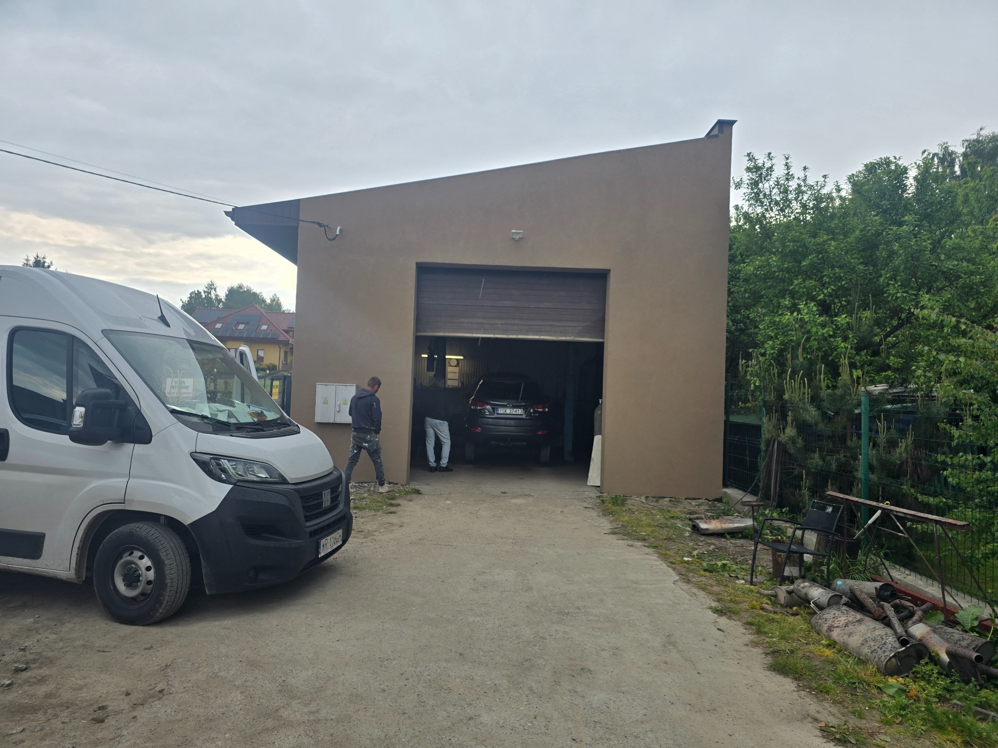 Beżowa elewacja budynku z otwartą bramą garażową, widoczny biały bus i osoby w środku. Uwagę zwracają rury wydechowe leżące obok ścieżki.