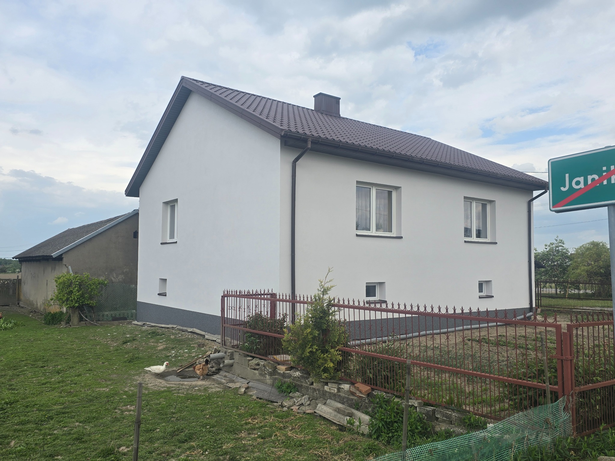Biały dom z brązowym dachem po renowacji elewacji, widoczny zza czerwonego ogrodzenia, z tablicą miejscowości w tle i budynkiem gospodarczym z lewej.