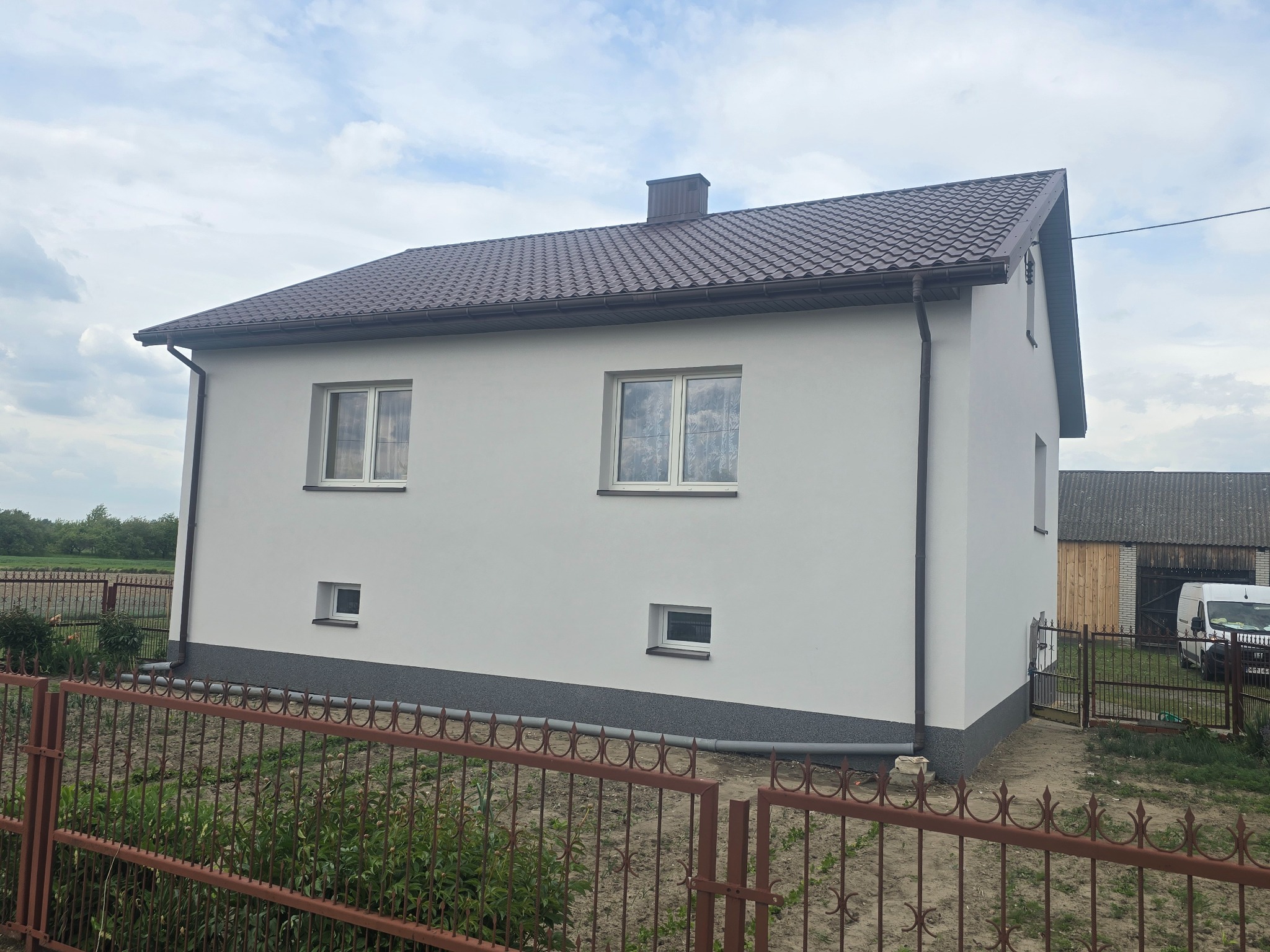 Dwukondygnacyjny dom z ocieploną, jasną elewacją i brązowym dachem z blachodachówki, widziany zza metalowego płotu. Widoczne okna i fragment posesji.