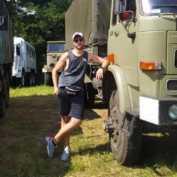 Mężczyzna w sportowym stroju opiera się o koło wojskowej ciężar&oacute;wki Star 244 na tle zieleni. Widoczne inne pojazdy wojskowe.