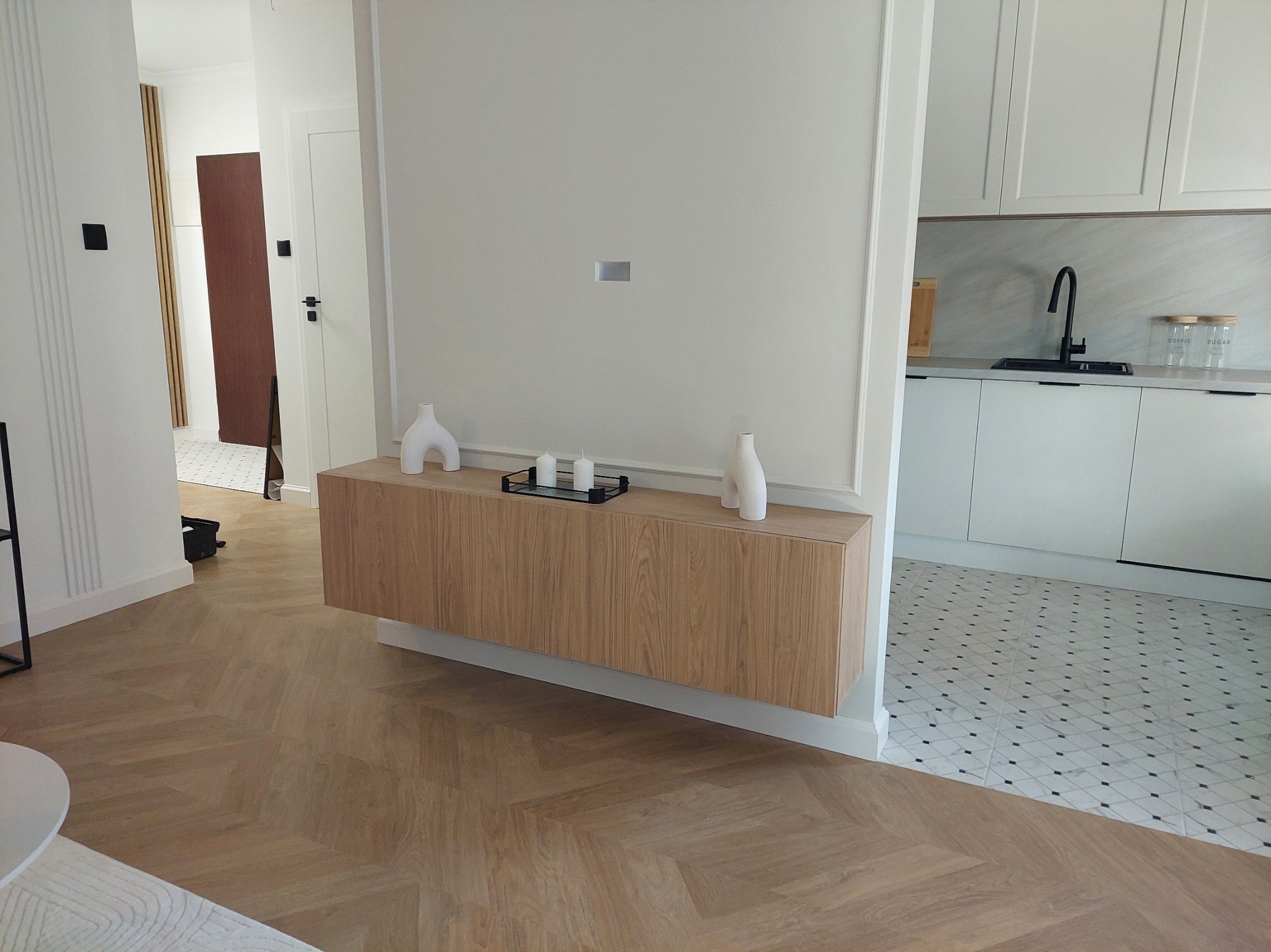 Nowoczesna szafka RTV z drewnianymi frontami, wisząca na ścianie w salonie, z dekoracjami. W tle widoczny fragment kuchni z białymi szafkami.