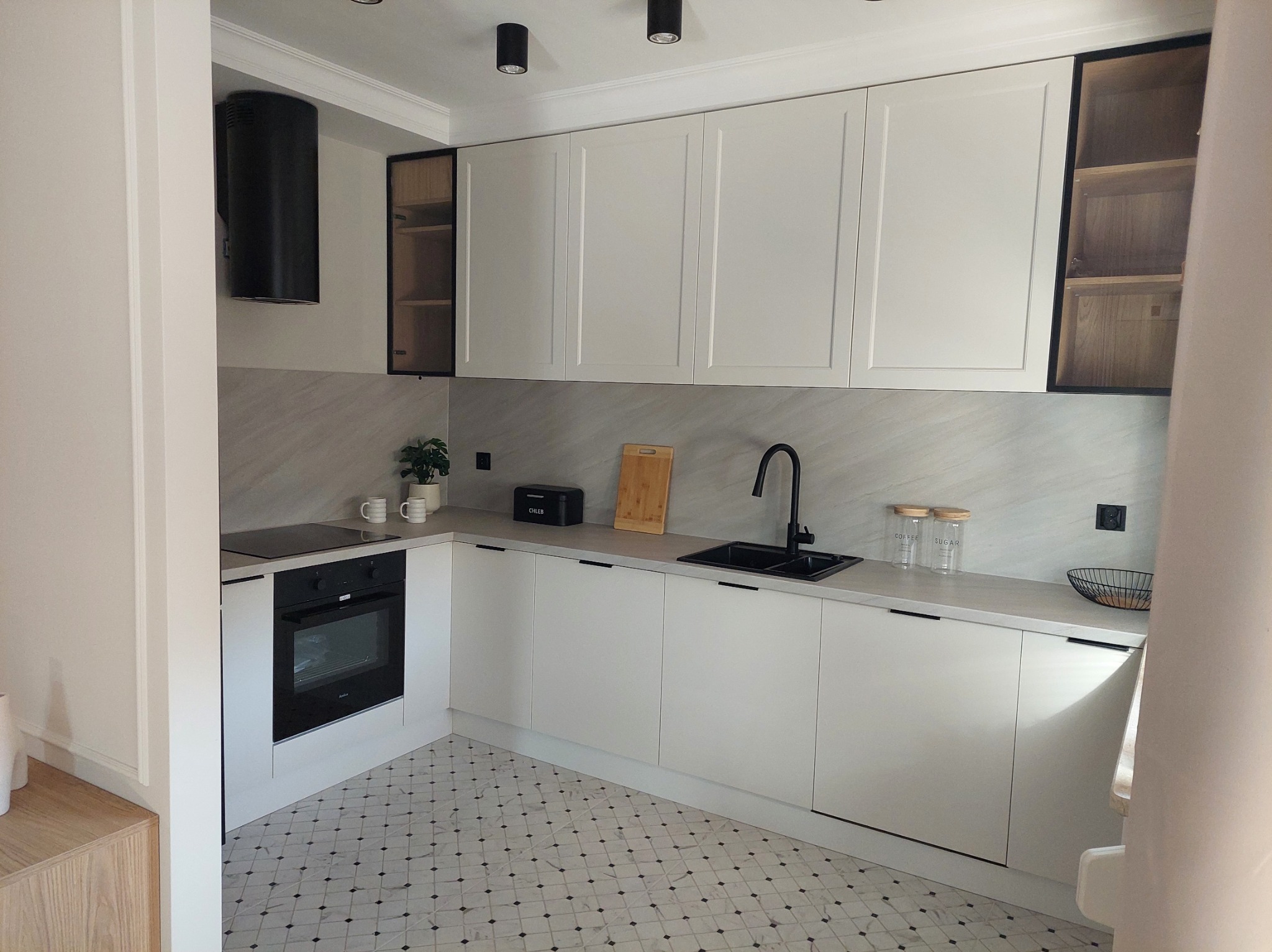 Nowoczesna kuchnia z białymi frontami i czarnymi akcentami, widok na narożną zabudowę z piekarnikiem, płytą i zlewozmywakiem, wzorzysta podłoga.